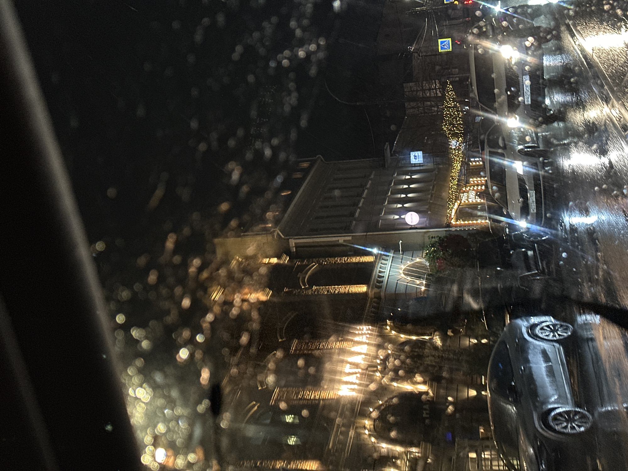 The image shows a nighttime street scene in Kuleshovka, Russia, likely taken from inside a vehicle during wet weather. Raindrops obscure the view. A building with illuminated steps and decorative lights is visible, along with a Christmas tree. Cars are parked along the street. Streetlights illuminate the scene. A streetlight with a pedestrian crossing sign can be seen on the corner of the street.