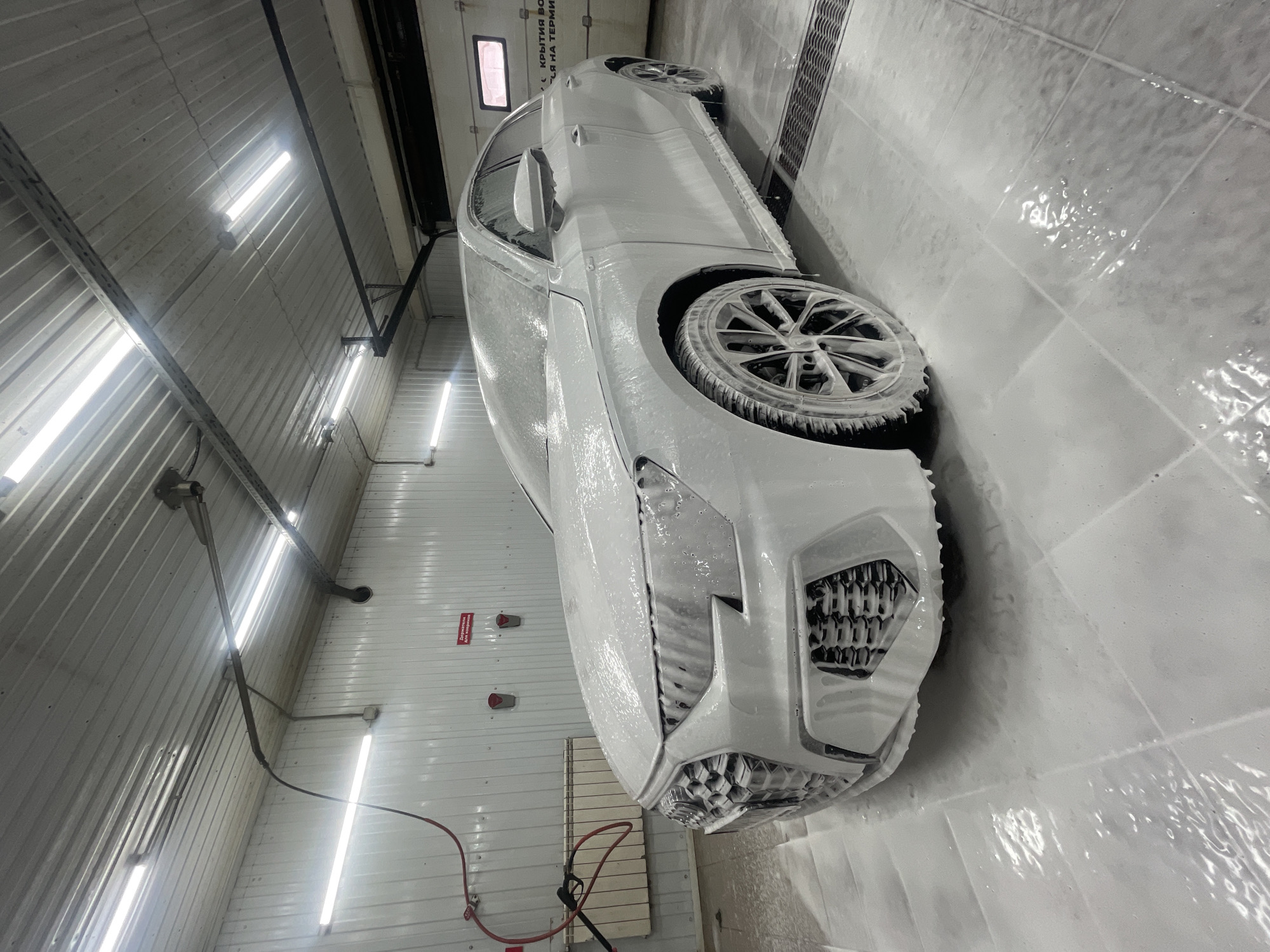 The image depicts a white car, heavily coated in white foam, seemingly undergoing a car wash. The car is inside a garage-like structure with a white tiled floor, and the walls are also white, possibly corrugated metal. Fluorescent tube lights are visible on the ceiling. There are various warning signs written in Russian on the walls. A red hose is visible near the bottom left corner. The location appears to be a car wash facility in Krasnoyarsk, Russia, based on the Cyrillic text on the signs.