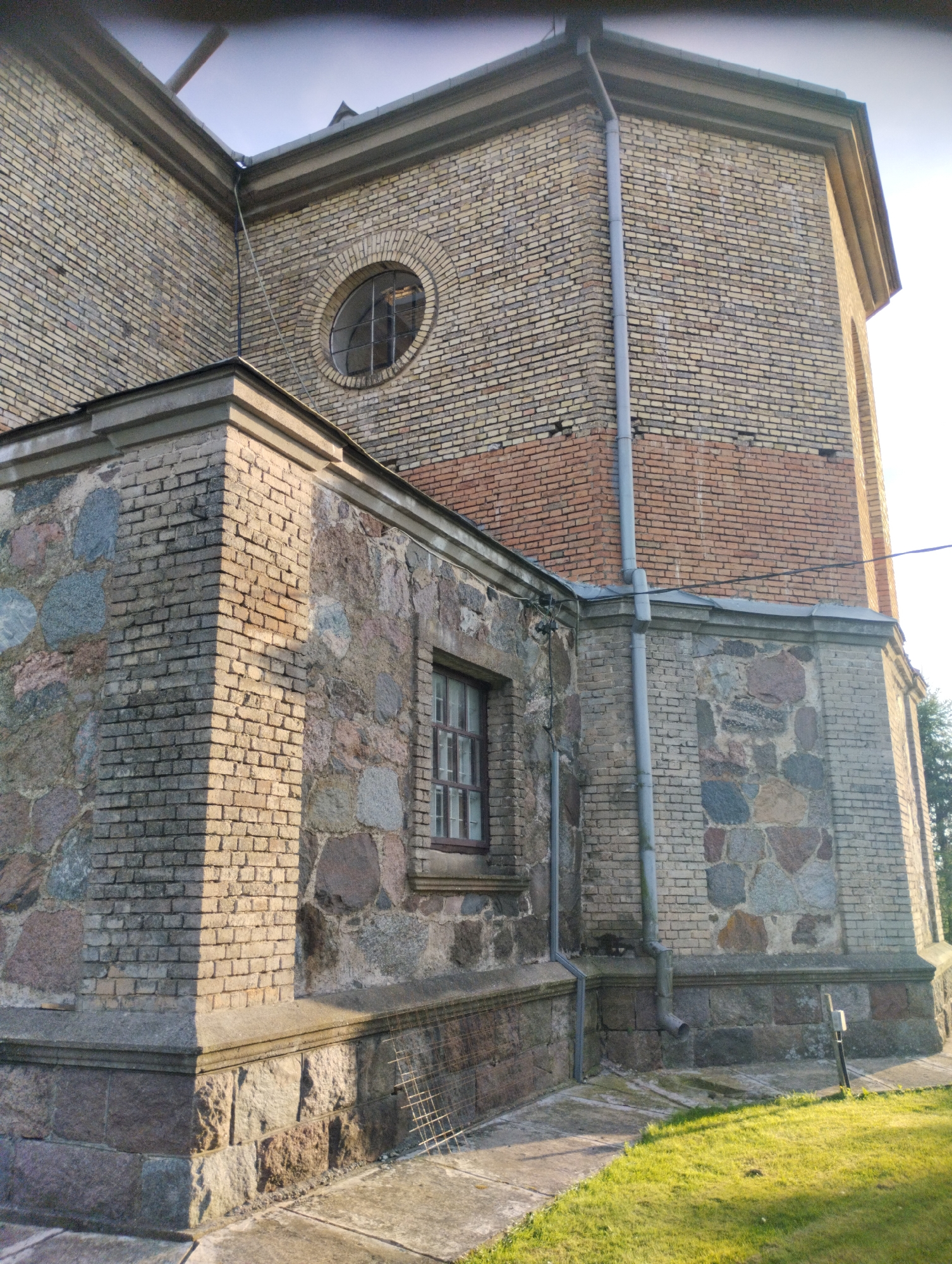 The image shows the corner of a brick building with large stone blocks on the lower section of the walls. A drainpipe runs down the side of the building. There is a round window high up on the brick wall and a rectangular window on the stone section. The ground is a mixture of concrete paving stones and green grass. The building is located in the city of Vishnyovy Sel's'kiy Sovet, Belarus.
