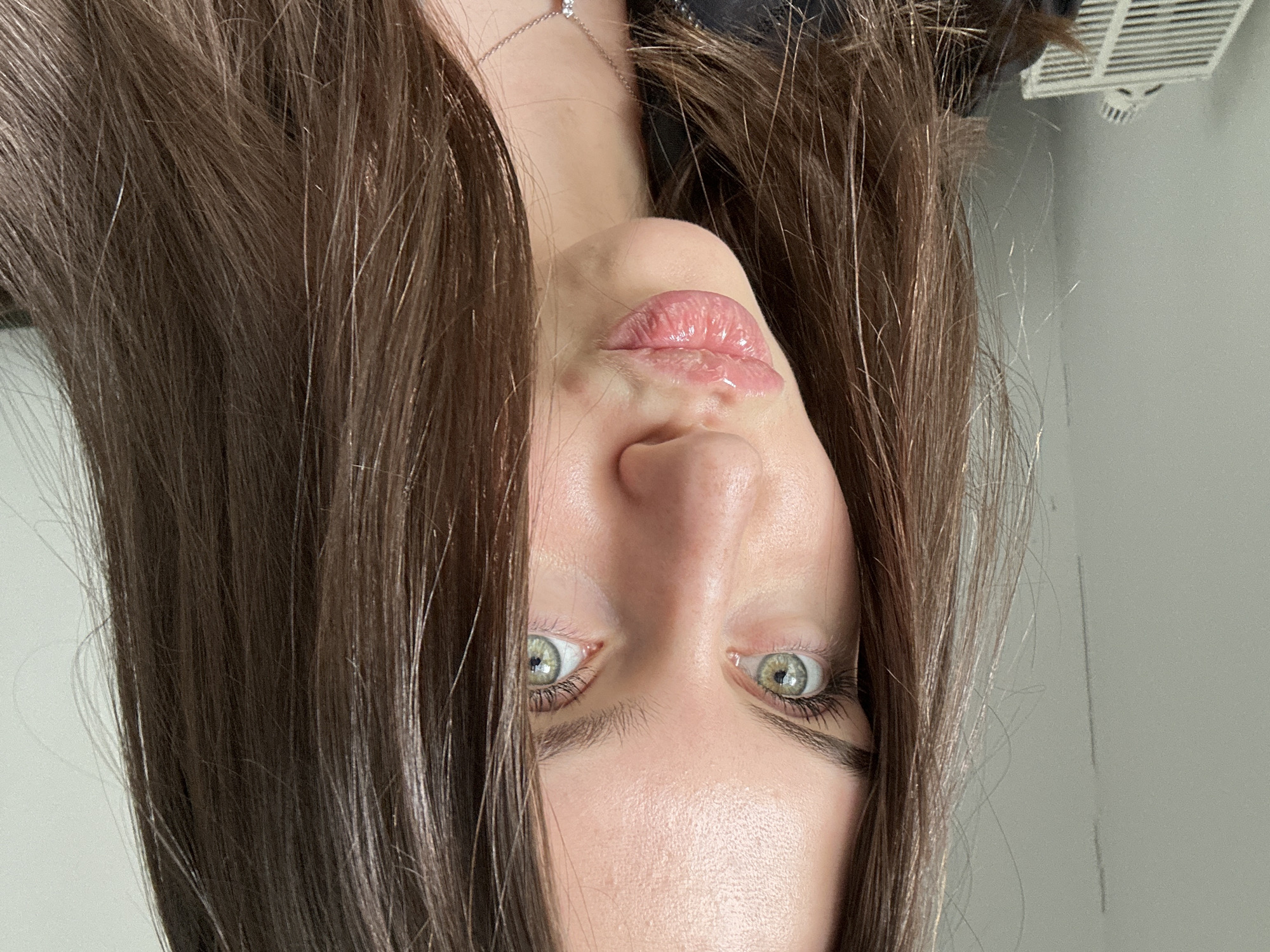 A portrait of a woman with fair skin and green eyes, with brown hair framing her face. Her lips are slightly pursed. A silver necklace is visible on her neck. The background is a gray wall with a white ventilation panel in the top right corner. The location is Unknown, Unknown.