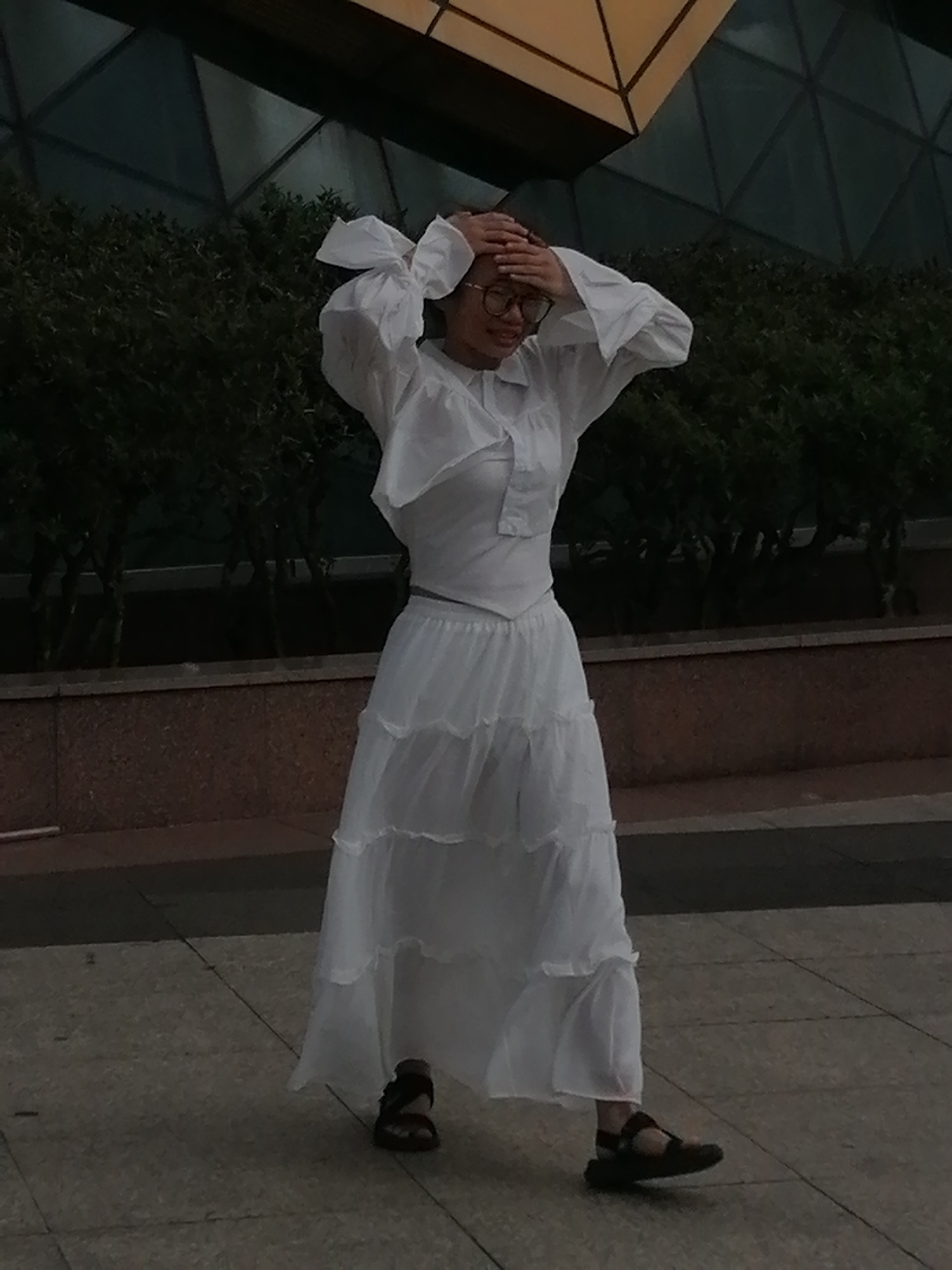 A young woman with glasses stands outdoors in Da Lat, Vietnam, her hands clasped on her head as she looks down and slightly to her right. She is wearing a white tiered dress with a ruffled collar and long, voluminous sleeves, along with dark sandals. The background features lush green bushes and a building with a geometric, angular facade. The ground is paved with square tiles.