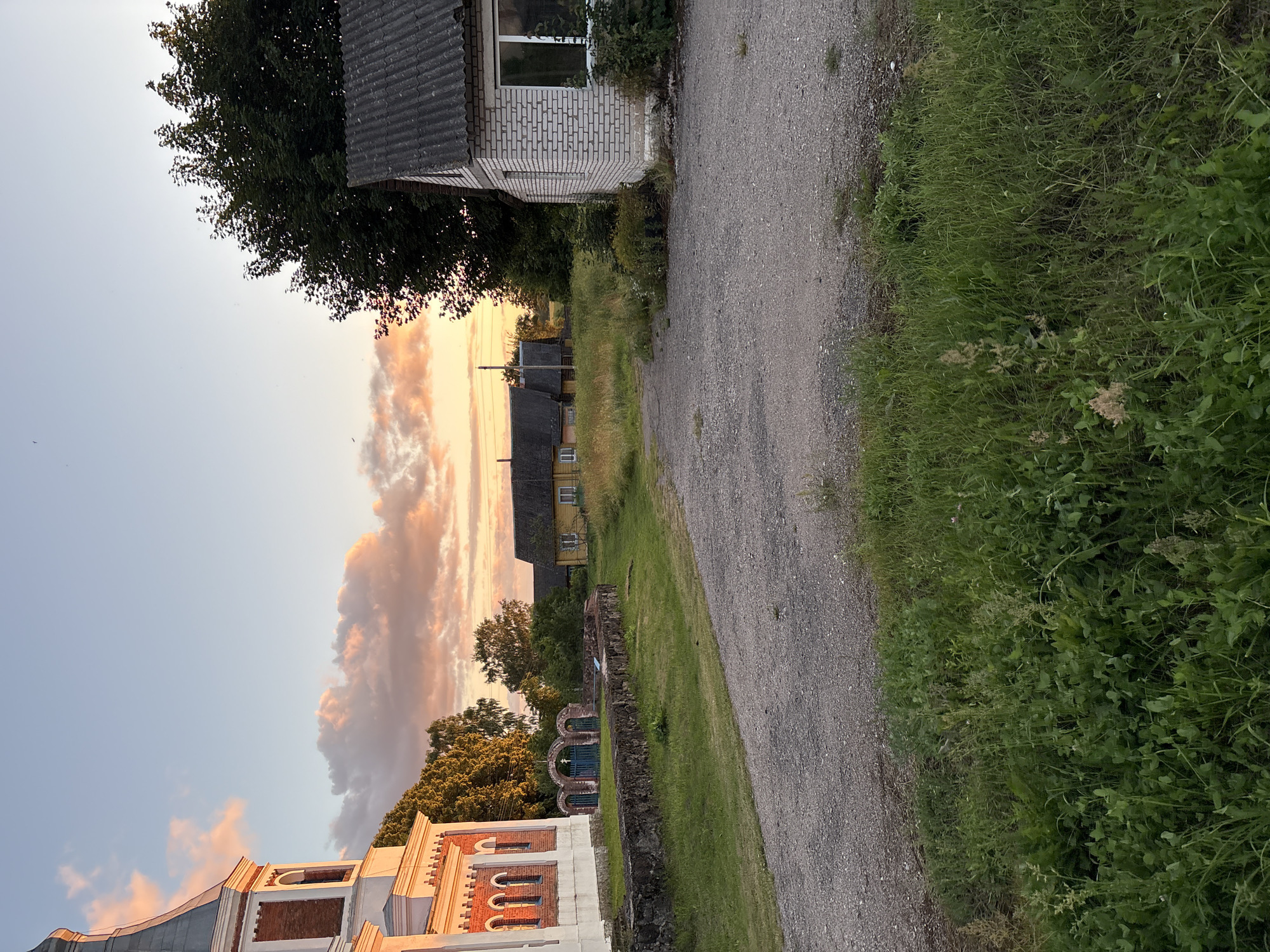 The image depicts a rural scene at dusk, with a warm, golden light illuminating the sky and casting long shadows. In the foreground, a gravel path leads towards the center of the frame. To the left, a portion of a red and white brick building, likely a church due to its architectural style and steeple, is visible. Adjacent to it is a low stone wall with an arched, green metal gate. In the middle ground, a long, yellow wooden house with a dark roof stands among tall green grass and trees. To the right, a small, white brick building with a grey, corrugated roof and a large window is partially obscured by lush vegetation. The overall environment suggests a quiet, possibly abandoned or historical, rural settlement in Belarus, specifically within the Відзаўскі сельскі Савет. The sky is a pale blue with streaks of orange and pink clouds, indicative of sunset. No people or animals are visible in the image.