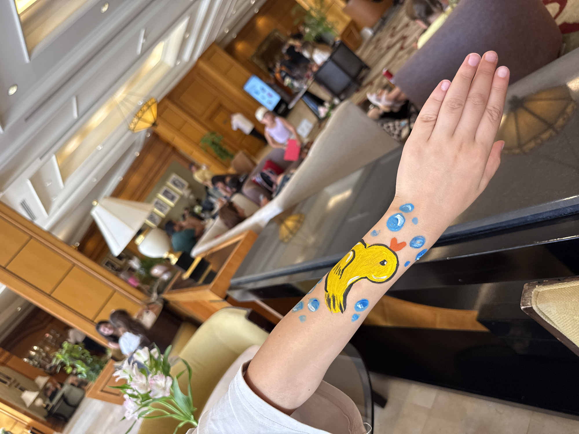 The image shows a close-up of a person's arm with a yellow duck and blue bubbles painted on it, suggesting a temporary body art. The background is a blurry interior of what appears to be a hotel lobby or lounge area. There are people visible in the background, some seated and others standing, with furniture like sofas and chairs. Light fixtures are visible on the ceiling, and there are decorative elements such as plants and framed pictures on the walls. The overall impression is a casual, indoor setting. The mention of Sharm El Sheikh, Egypt, is not supported by any visual elements in the image.