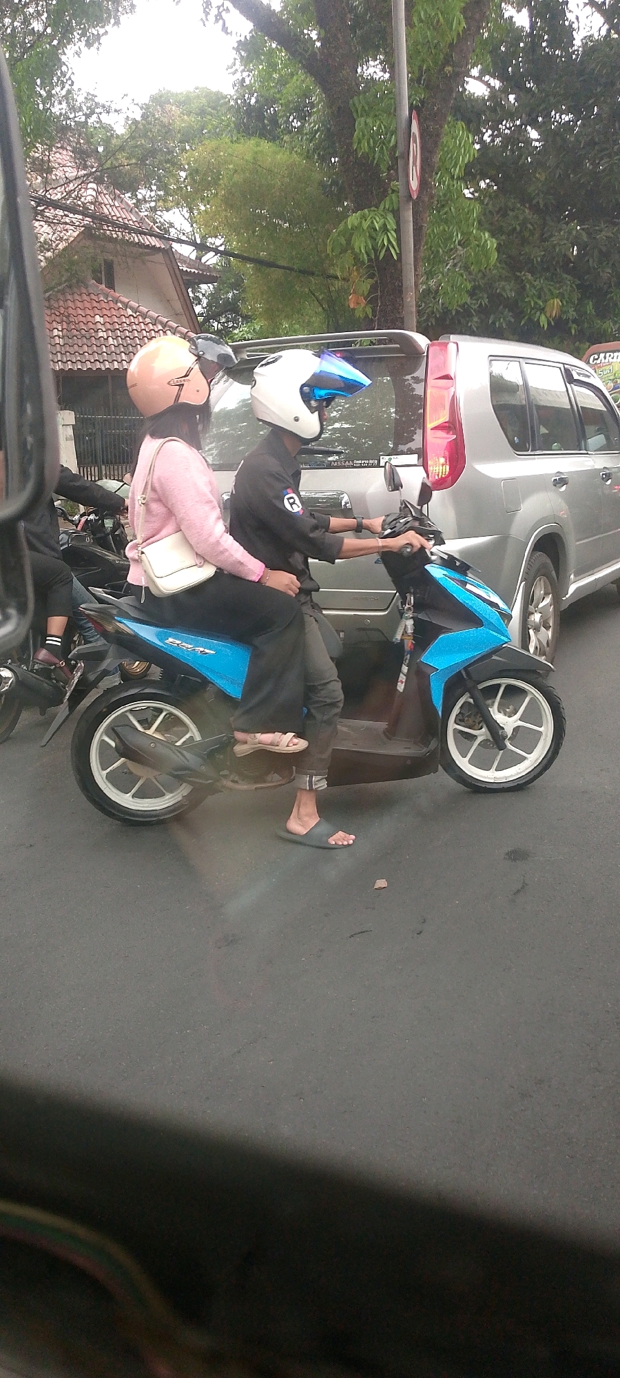 Two individuals are riding a blue scooter on an asphalt road, likely in the city of Bandung, Indonesia, based on the architectural style of the nearby building and lush foliage. The person in the back, wearing a light pink sweater and black pants, has a beige handbag slung over their shoulder and a light brown helmet. The person in the front, wearing a black shirt with a white "R" logo and dark pants, is wearing a white helmet with a blue visor. The scooter has a blue body and white wheels. In the background, a silver SUV is visible, as well as a building with a terracotta tiled roof and dense green trees. The lighting suggests it is daytime, possibly overcast.
