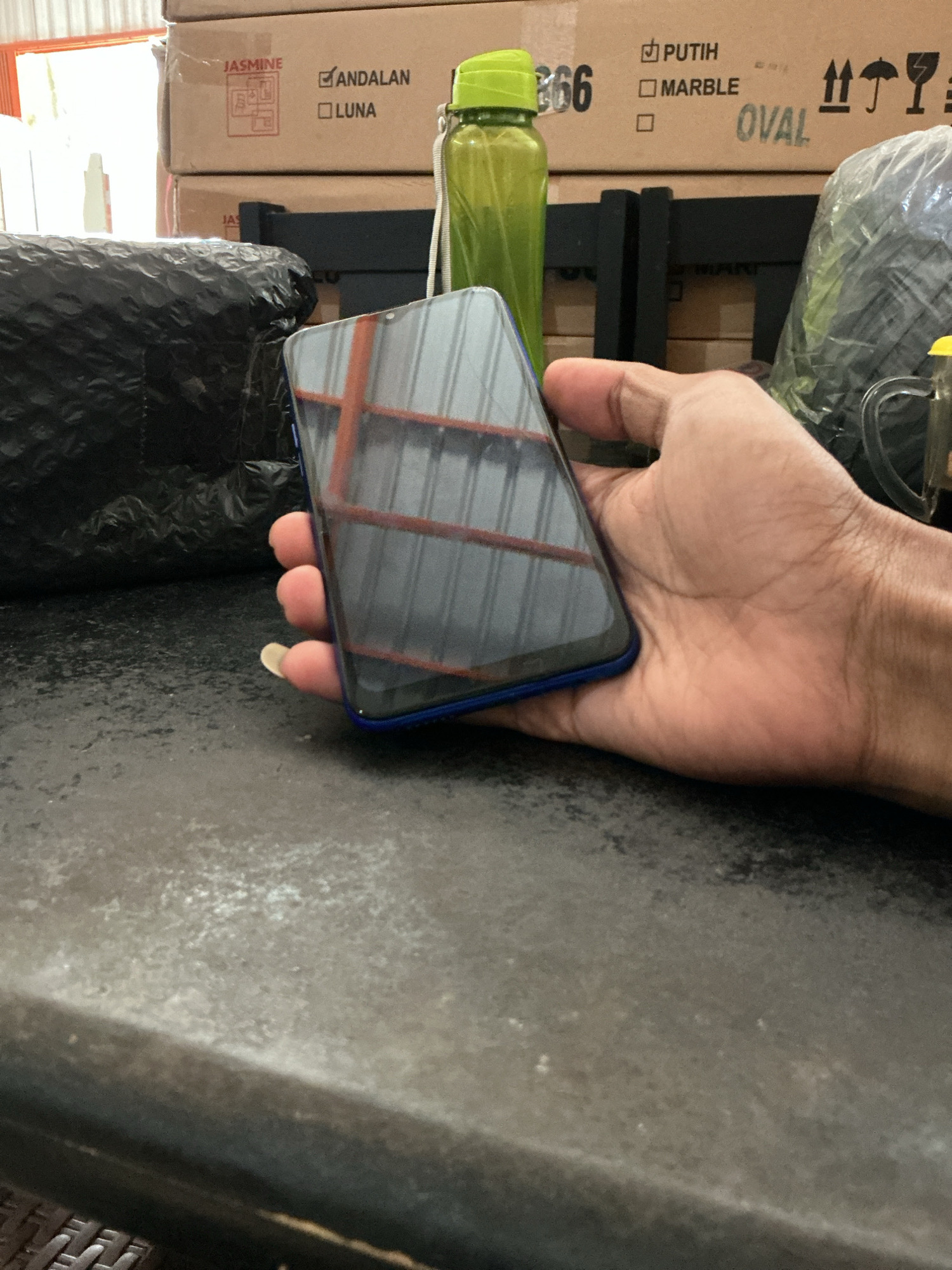 A person's hand is holding a black smartphone with a blue frame. The phone's screen reflects a rack of metal shelves. In the background, stacked cardboard boxes are visible. One box has the text "JASMINE," "ANDALAN," and "LUNA" printed on it. Another box has the text "PUTIH" and "MARBLE" printed on it, along with the word "OVAL." A green plastic water bottle is positioned behind the phone. The scene appears to be indoors, possibly in a storage area or warehouse, given the presence of boxes and industrial-looking shelving. The surface in the foreground is a dark, textured countertop. The overall lighting suggests an indoor setting with overhead illumination.