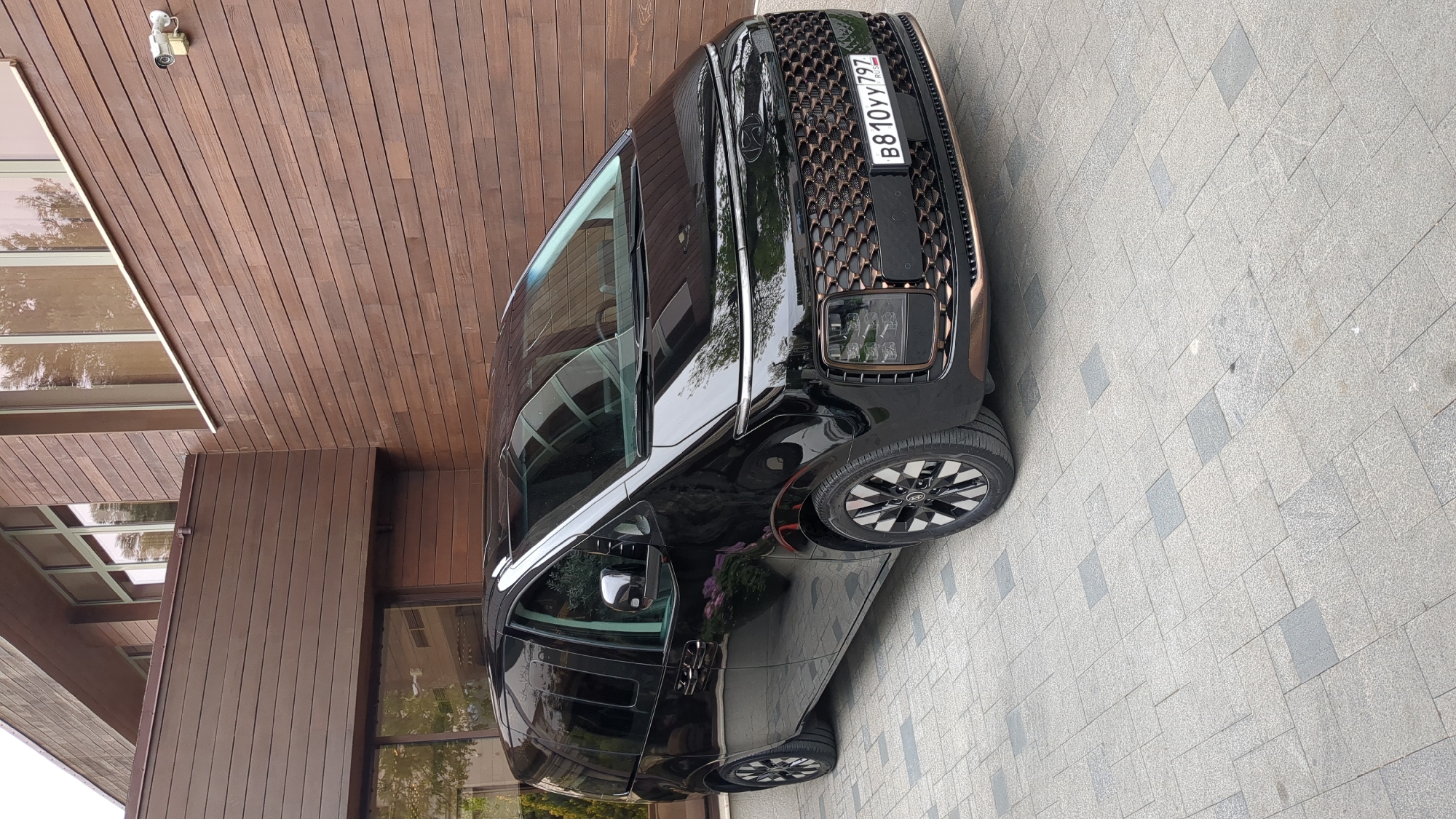 The image shows a black Kia EV9 car parked on a paved surface next to a building with brown wooden cladding and large windows. The car has a Russian license plate B810YY797. The location is likely Gelendzhik, Russia, based on the information in the filename. A security camera is mounted on the wooden cladding above the building's windows. The day appears to be overcast.