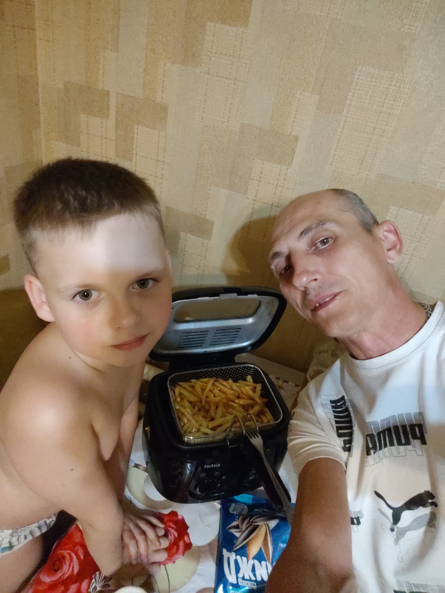 This media file depicts two individuals, a young boy and an adult man, in what appears to be an indoor setting. The boy, in the foreground and to the left, has light brown hair and is looking directly at the camera with a neutral expression. He is shirtless and wearing patterned undergarments. Next to him and slightly behind, the adult man, with a shaved head and fair skin, is also looking towards the camera. He is wearing a white t-shirt with Puma branding.
Between them is a black deep fryer filled with golden french fries. A silver fork rests on the edge of the fryer basket. To the lower right, a blue bag with images of sunflower seeds and text is visible, and to the lower left, a red cloth with a floral pattern is partially seen. The background features a textured wallpaper in shades of beige and yellow. The lighting suggests it is daytime, possibly in the evening given the indoor setting and artificial light. There are no clear indications of weather or specific time of day beyond it being a casual, domestic scene. The overall emotion conveyed is relaxed and informal.
Visible text includes "PUMA" on the man's shirt and partially obscured text on the blue bag which appears to be "ДЖУЖА PREMIUM" in Cyrillic. The fryer itself has some text on it, possibly a brand name, but it's illegible. The context provided indicates the location as Bor, Russia.