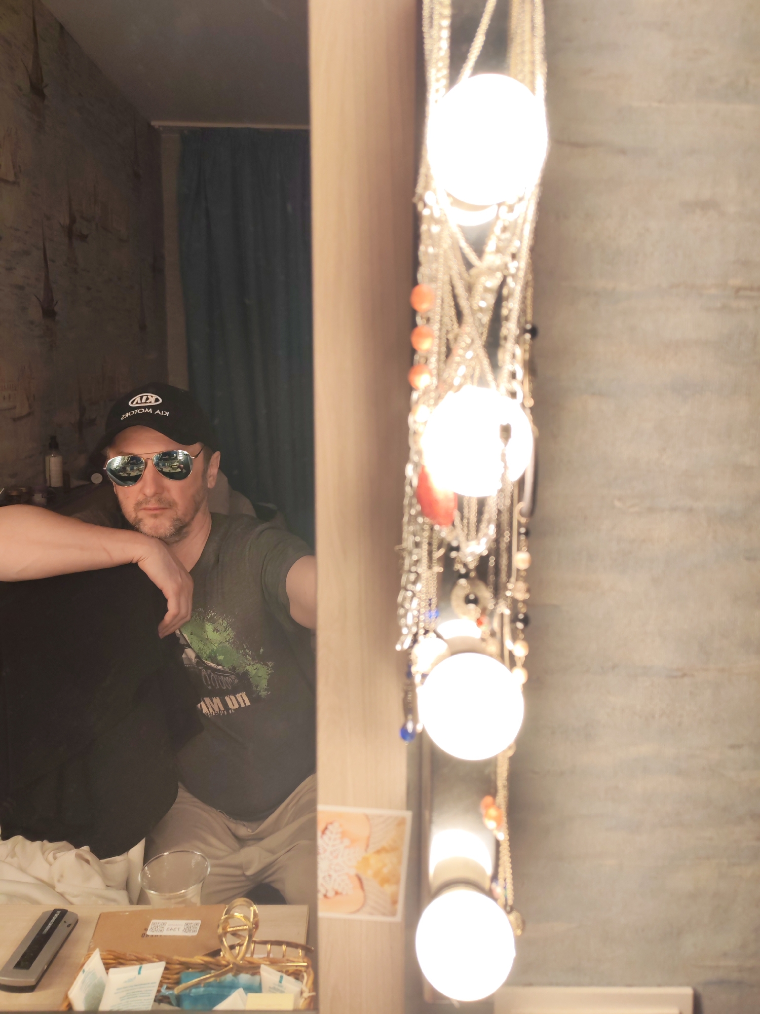 The image depicts a man with a goatee and sunglasses, wearing a black baseball cap, reflected in a mirror. The cap has the logo "KIA MOTORS" on it. The man is wearing a green t-shirt. The reflection shows a room with a curtained window, a patterned wall, and various items on a table. The mirror is surrounded by decorative hanging chains and exposed light bulbs. The table contains a remote control, a box, some small packets, and a water glass. There appears to be an image or postcard stuck to the side of the mirror. The image was taken indoors in the city of Chaikovsky, Russia.