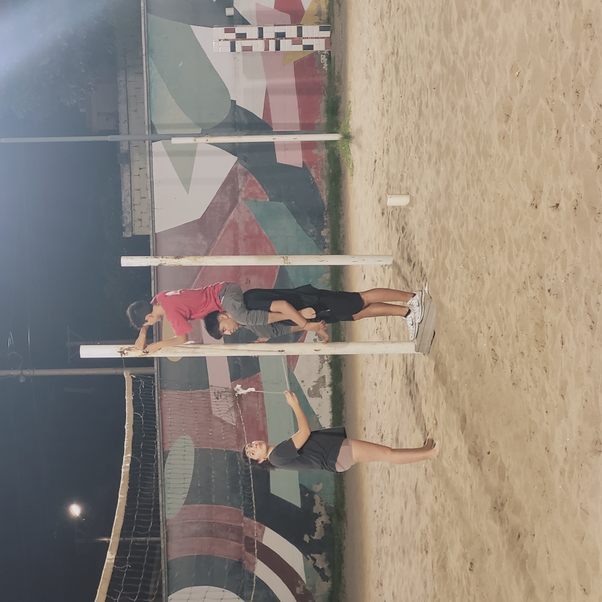 The image depicts a nighttime scene at a beach volleyball court in San Fernando del Valle de Catamarca, Argentina. The court is situated near a wall adorned with a geometric mural in shades of white, green, red, and black. Three people are present. A woman stands on the sandy court near the volleyball net, holding a white object. Two younger individuals are interacting on parallel bars. One youth, wearing a red shirt, is hanging onto the bar while another, wearing a gray shirt and black shorts, is positioned on top of them, creating a stacked arrangement. The ground is light-colored sand. A street light is visible in the background.