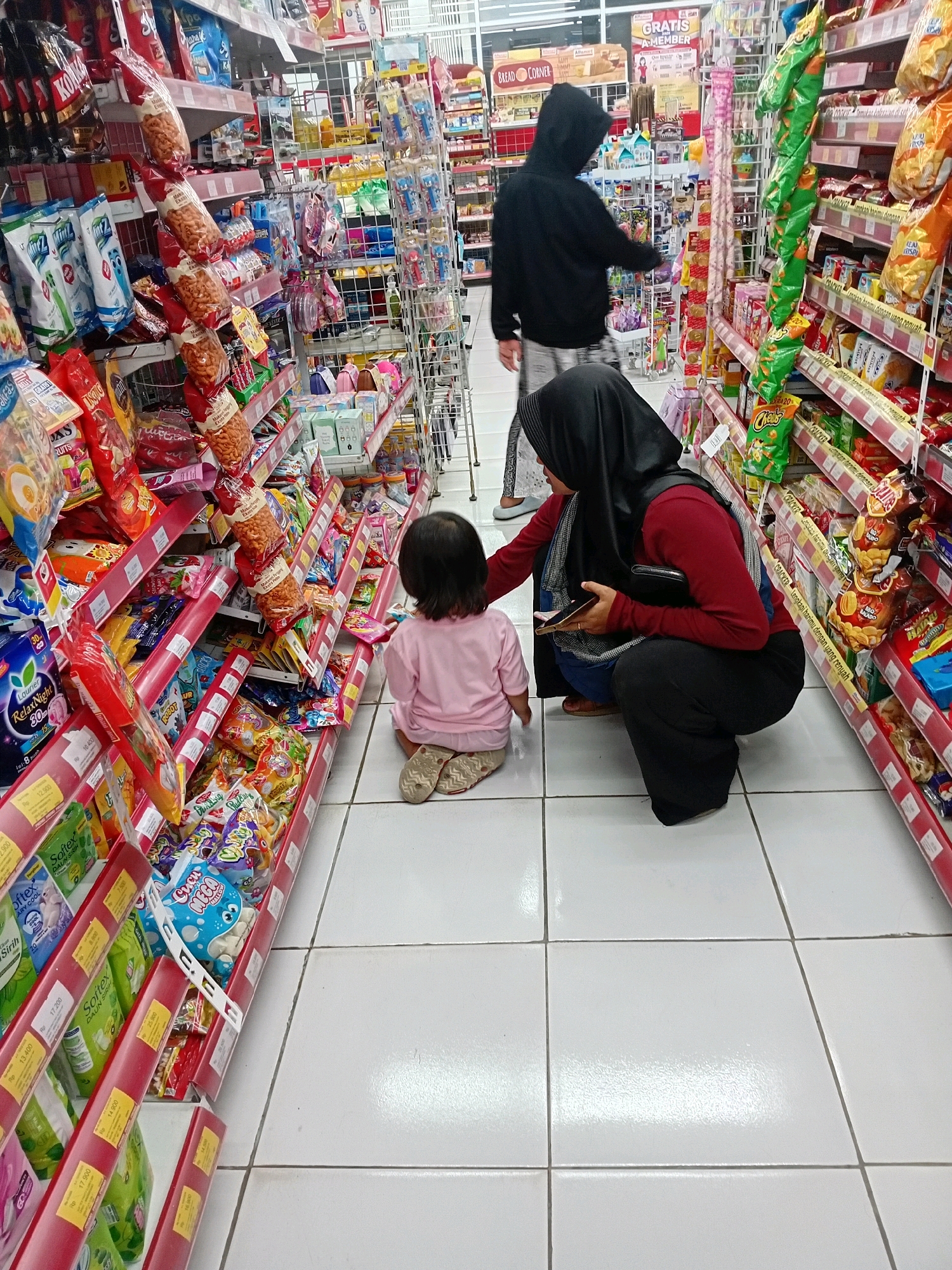 In Seren, Indonesia, a grocery store aisle is depicted with a woman in a black hijab and red shirt squatting next to a young child sitting on the floor. The woman is holding a smartphone and a wallet. Another person wearing a black hoodie is standing in the background, looking at the shelves. The aisle is lined with colorful products, including snacks, drinks, and toiletries, displayed on red shelves. The floor is tiled with white squares.