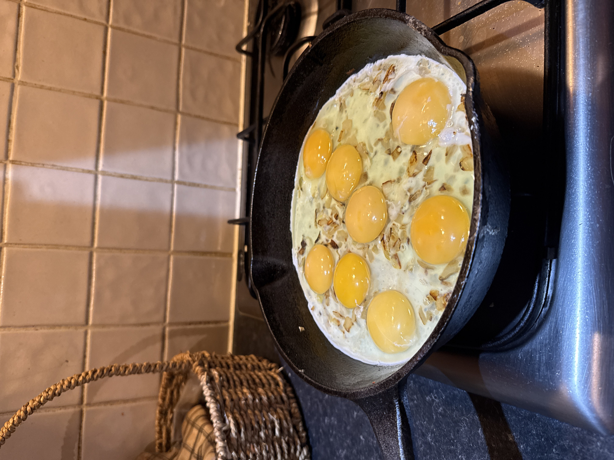 A cast iron skillet sits on a black stovetop next to a chrome utility sink, containing seven eggs cooking with small pieces of fried onion or garlic. The stovetop is part of a gas range, with black metal supports visible behind the skillet. A woven basket sits to the left of the stovetop, partially visible. The background is a wall with beige, square tiles with dark grout. The image does not depict any discernible features that would place it in Great Yarmouth, United Kingdom.