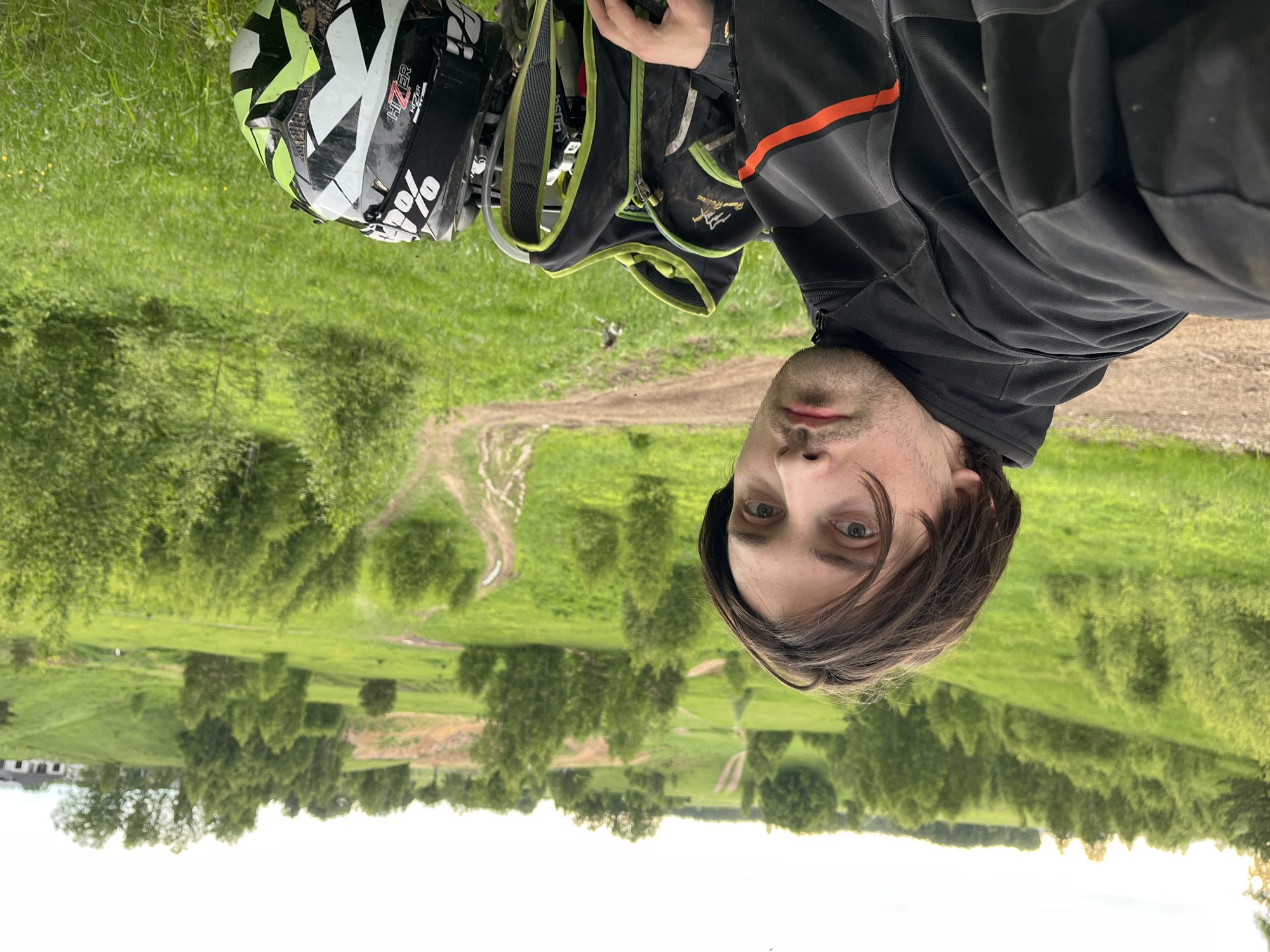 A man stands outside in Новочувашский, Russia, with a helmet and backpack. The man faces the viewer and appears to be taking a selfie. He has brown hair, blue eyes, and stubble. He wears a black jacket with an orange stripe on the shoulder. Next to him is a black and white helmet with green accents. The helmet has the brand name "HIZER" on it. Behind the helmet, a green backpack is hanging. The landscape consists of a green field with trees, and a dirt path can be seen in the distance. A building is visible in the far background.