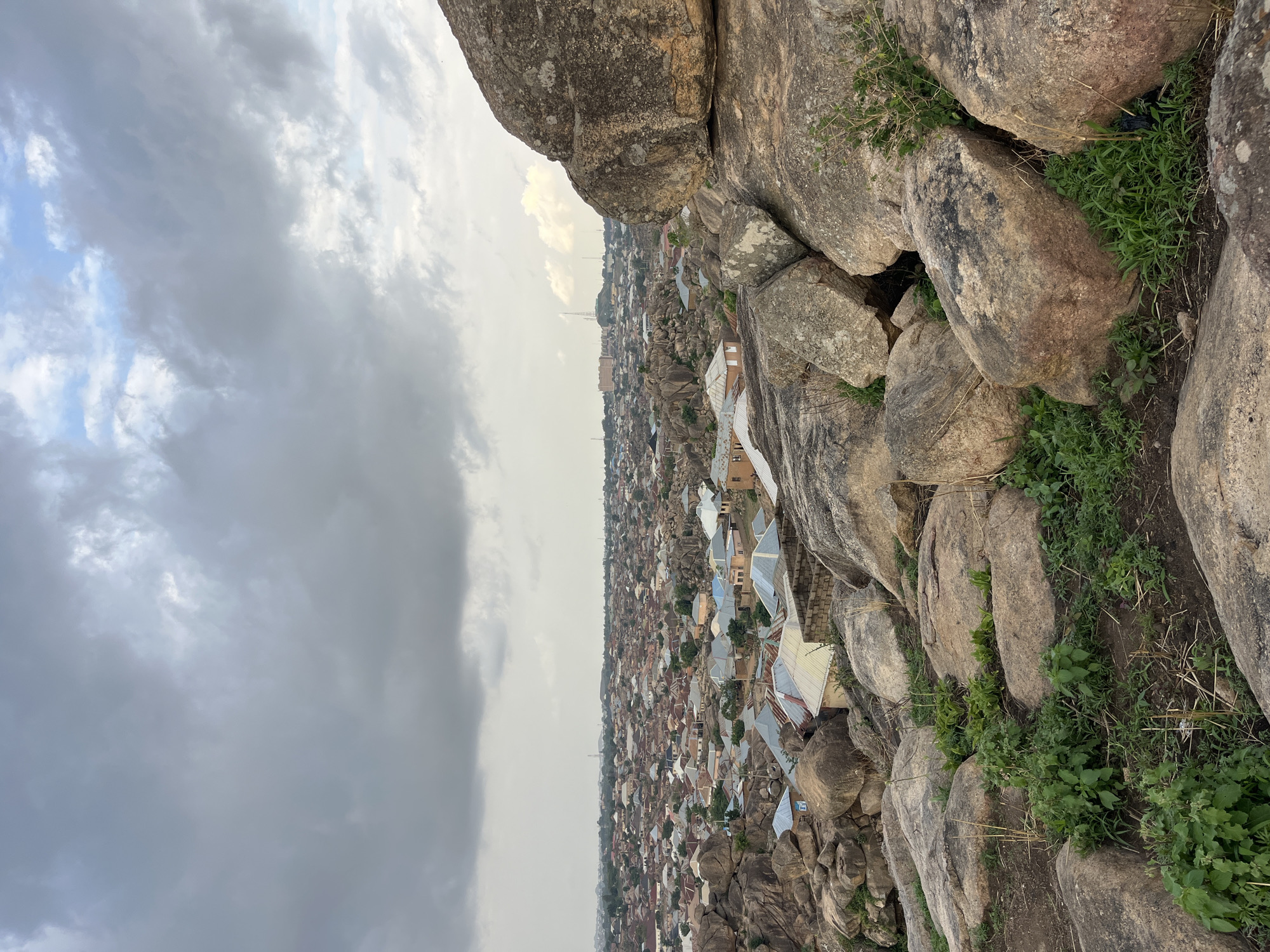 The image captures an elevated view overlooking a densely populated urban area, possibly in Jos, Nigeria, given the rocky terrain typical of the region. The foreground consists of large, irregularly shaped rocks and boulders, some of which are covered with patches of green vegetation. In the distance, the city sprawls out, with numerous buildings featuring corrugated metal roofs. The structures appear closely packed together, indicating a high population density. The sky is overcast with heavy, gray clouds. A tall communication tower or transmission structure is visible rising above the buildings in the middle ground.
