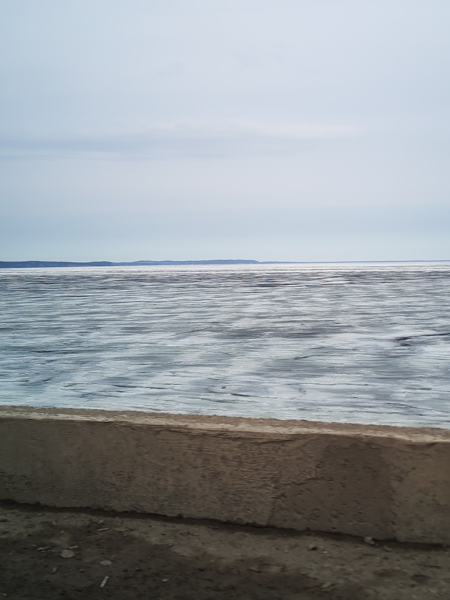 The image depicts a view of the Kama River in Chaikovsky, Russia, from a concrete embankment. The river is covered in thin sheets of ice with visible water underneath. The sky is overcast, with a light gray color. In the far distance, there is a thin strip of land, presumably the opposite bank of the river, with sparse vegetation. The foreground shows the top of the embankment, with small debris scattered around the base. There are no people visible in the image.