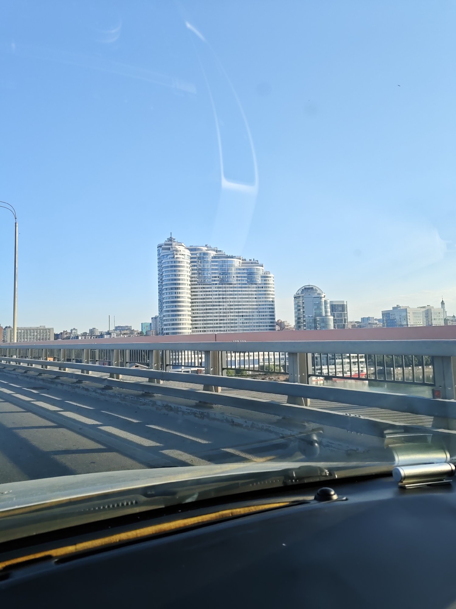 A sunlit, clear blue sky is visible over the city of Rostov-on-Don, Russia. The photograph is taken from a moving vehicle, likely a car, with the dashboard visible in the foreground. The view is from a bridge overlooking a cityscape. The most prominent structure is a distinctive, modern, wave-shaped white building with many windows. To the right of this building, there is a rounded, glass-fronted building. Further in the distance, other buildings of varying heights and styles can be seen. A gray metal railing with horizontal bars separates the bridge's road from the scene below. Shadows from the bridge's structure are cast across the road. The light suggests it is either morning or late afternoon.