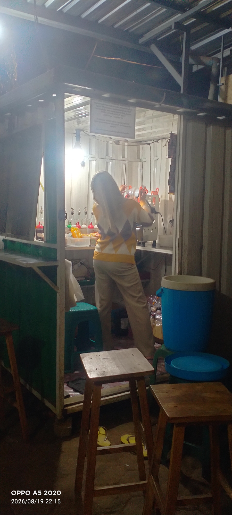 A person wearing a hijab and a yellow and grey patterned sweater is standing behind a food stall. The stall has a counter and shelves with various bottles and containers. In the background, a large blue cooler is visible. The scene appears to be outdoors, possibly a market or street food vendor, with a corrugated metal roof overhead. The lighting suggests it is evening or nighttime. The location is identified as Kedung, Indonesia.