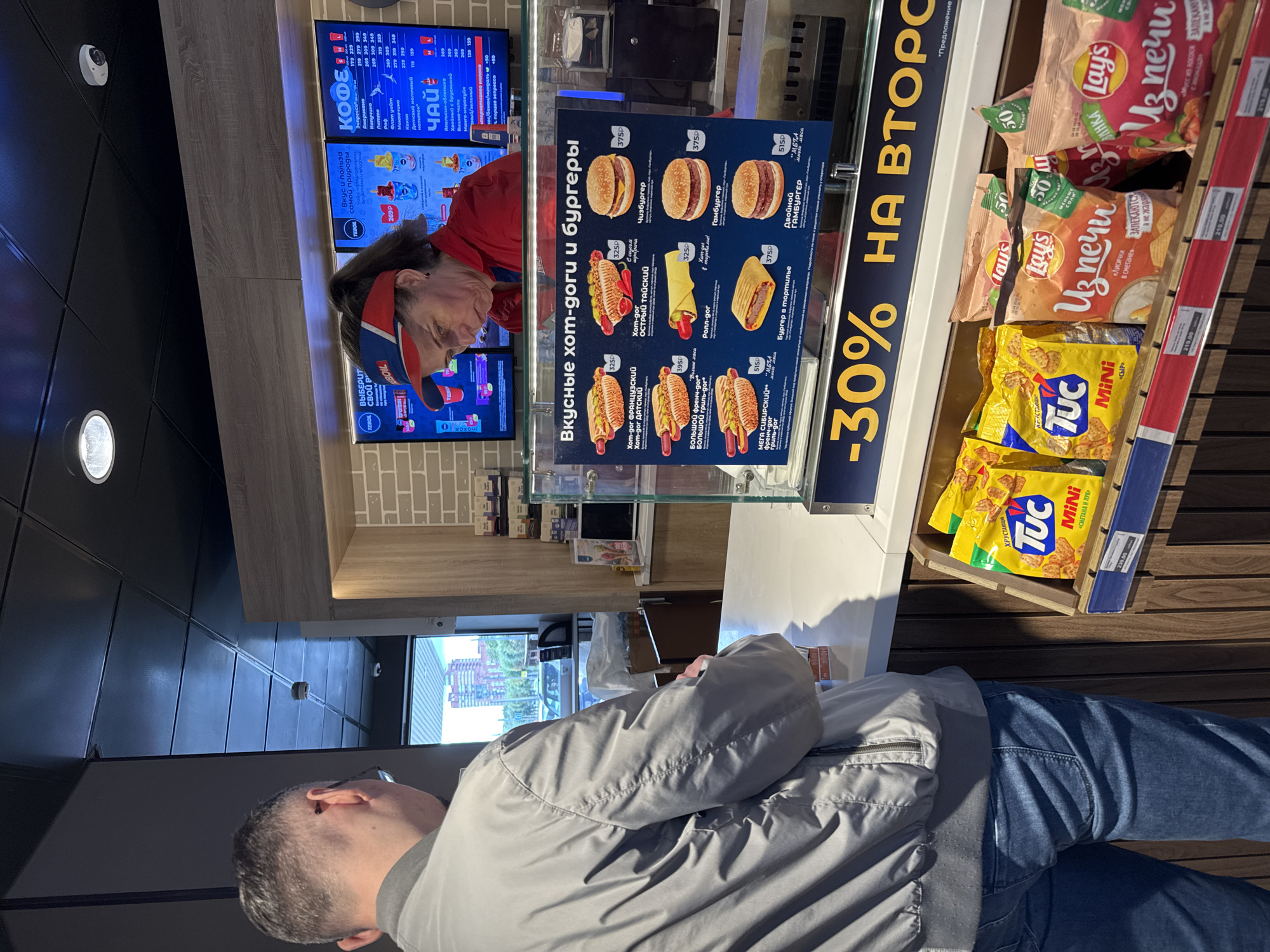 A man in a gray jacket and blue jeans stands in front of a counter in a convenience store. Behind the counter, a woman wearing a red uniform and a red visor is serving customers. The store appears to be a part of a gas station, with a large window in the background showing a gas station canopy and a street with buildings. The counter displays a menu of hot dogs and burgers, as well as various bags of chips and snacks. The overall scene depicts a typical transaction at a convenience store, likely in Saint Petersburg, Russia, given the Cyrillic text on the menu and product packaging.