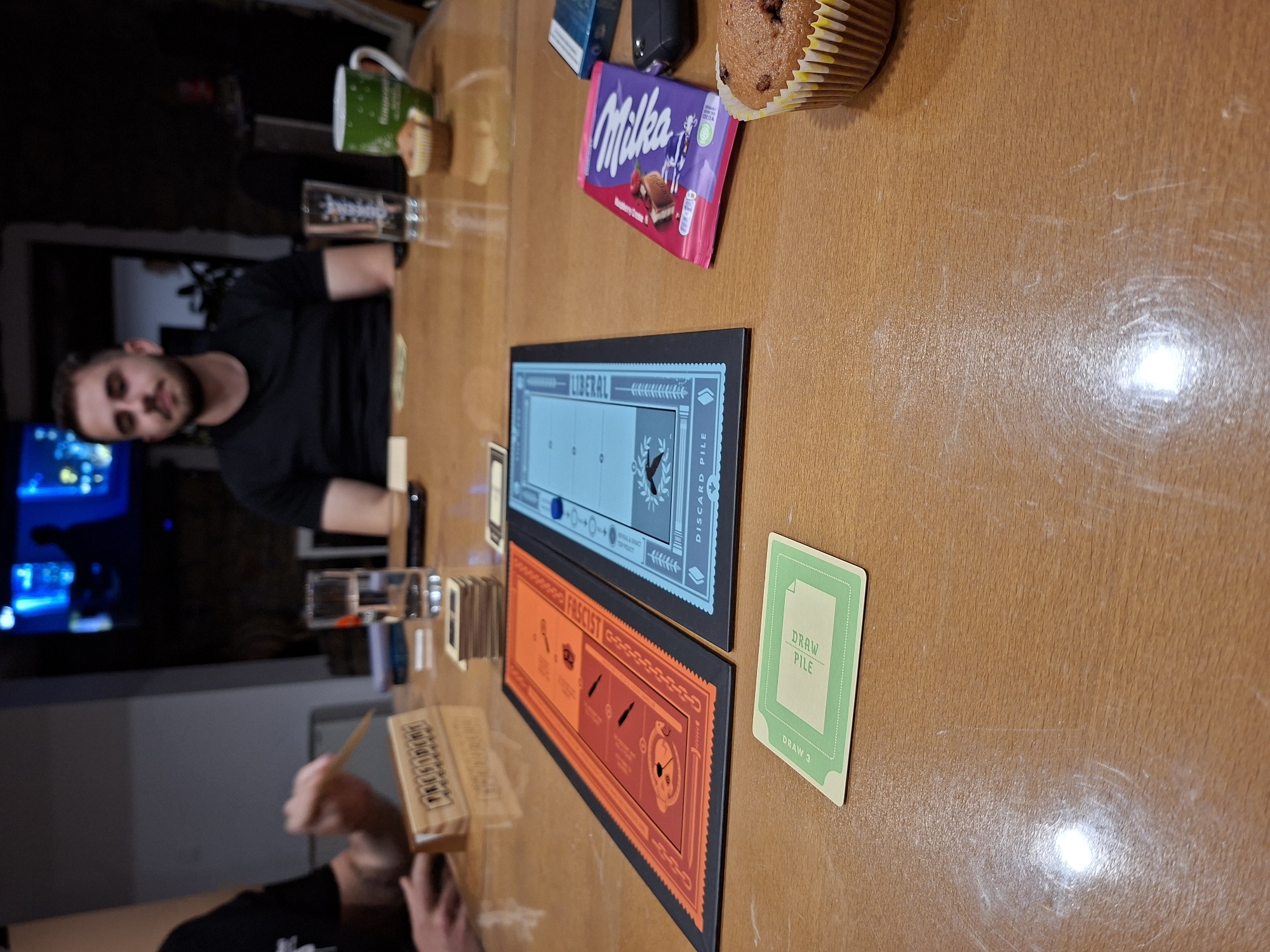 The image shows a tabletop scene. A man with a beard is partially visible on the left. A wooden table displays two game boards: a blue one labeled "LIBERAL" and an orange one labeled "FASCIST." A green "DRAW PILE" card is next to the boards. A wooden box with "PRESIDENT" printed on it is near the bottom-left corner. Other objects on the table include a Milka chocolate bar and a cupcake. The environment appears to be indoors. There is no verifiable evidence of Gabrovo, Bulgaria.