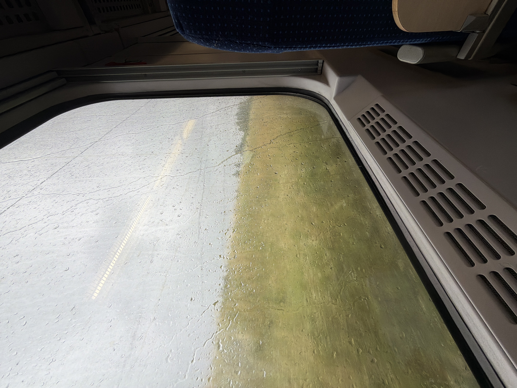 This media file shows a view from inside a train compartment looking out a rain-streaked window. The window is covered in numerous water droplets, obscuring the view of the outside landscape. The outside appears to be a mix of overcast sky and a greenish-brown field, suggesting a rural or semi-rural setting. A faint, striped reflection is visible on the left side of the window, possibly from the train's interior.
The interior of the train compartment is partially visible. In the upper part of the image, a blue patterned seat is seen, along with some overhead luggage racks. To the right of the window, a beige-colored vent with horizontal slits is present. The overall impression is a dreary, rainy day, likely in the daytime, with the view from the train conveying a sense of travel. No people are visible in the frame. There is no visible text. The location context of Żurawica, Poland, indicates a travel scenario within that region.