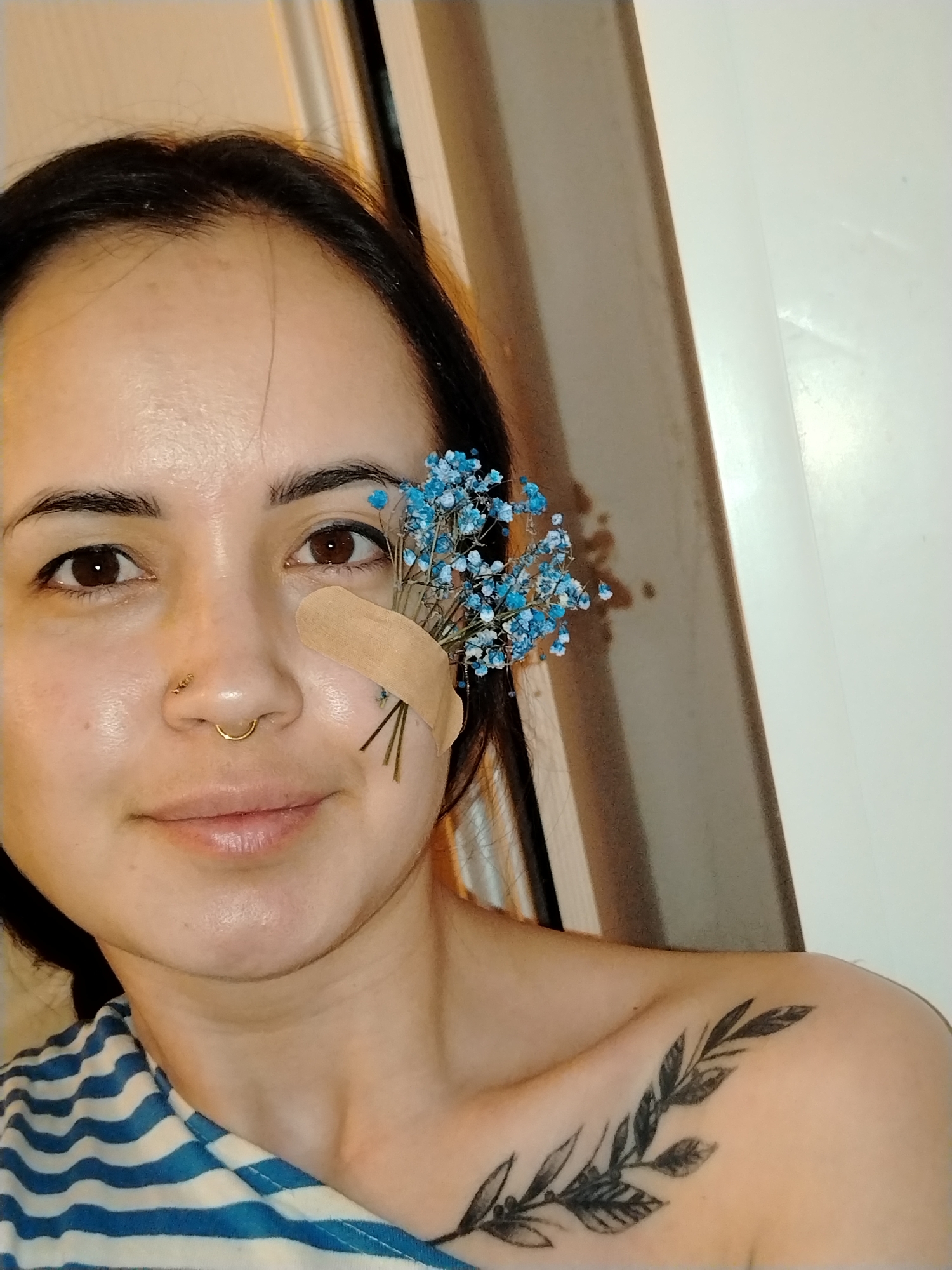 A woman is in a room, possibly a bathroom. She has dark hair and brown eyes, and is wearing a blue and white striped top. She has a bandage on her left cheek, and several sprigs of blue baby's breath flowers attached to the bandage. She has a gold nose ring in her left nostril. There is a plant tattoo visible on her right shoulder. Behind her is a white door and a brown doorframe. There is no information to indicate the location is Noviy, Russia.