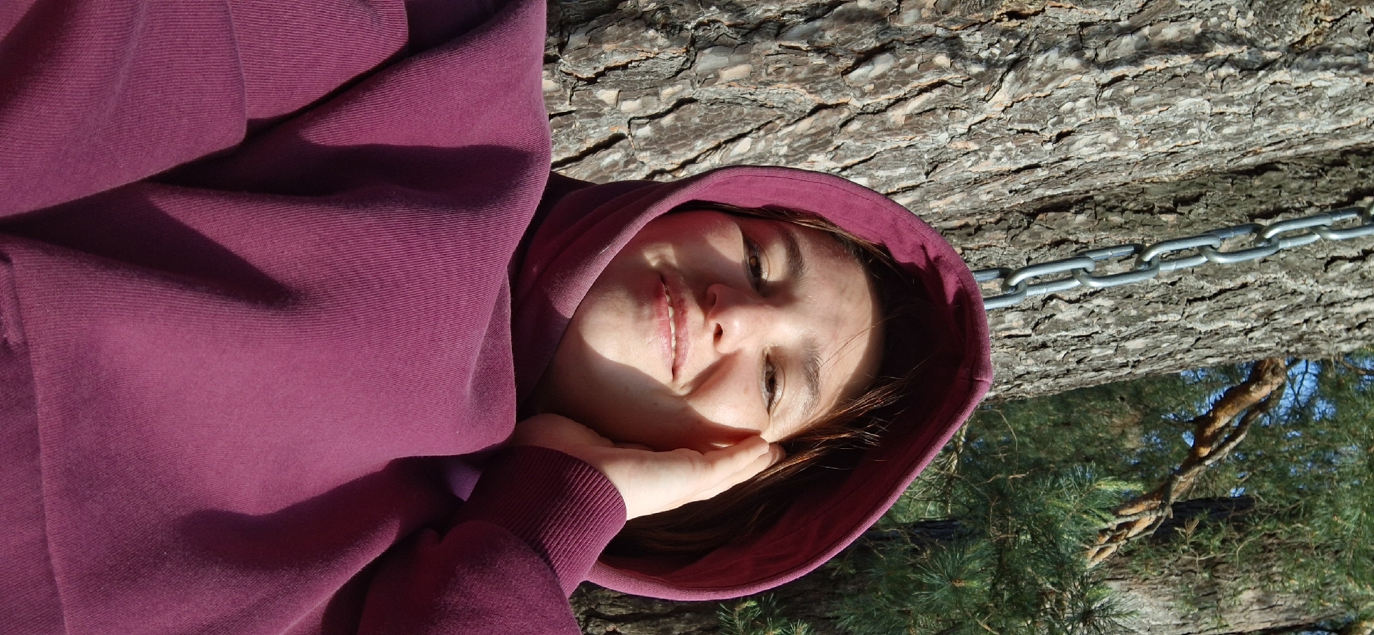 A young person wearing a maroon hooded sweatshirt is resting their head on their hand, near the bark of a tree, outside in the daytime. The photograph may have been taken in a park or wooded area within the city of Прикамский, Russia, as trees and a metal chain are visible in the background, which are consistent with an outdoor setting.