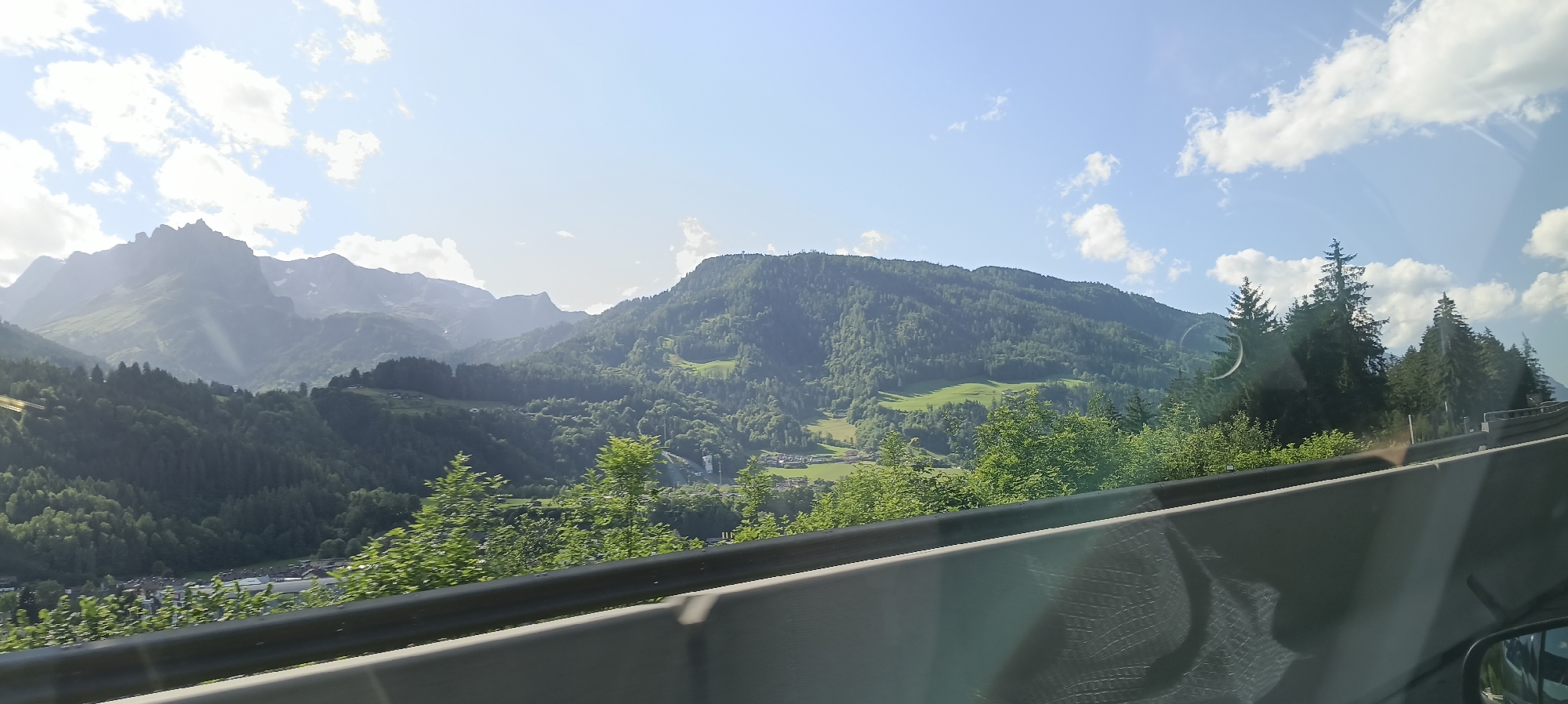 The image depicts a scenic view of mountains and a valley, likely taken from a moving vehicle. In the foreground, a concrete barrier with a metal railing runs horizontally across the frame, suggesting the viewpoint is from a road. The barrier is grey and textured, with a curved shape. Beyond the barrier, a lush green landscape unfolds, characterized by rolling hills covered in dense forestation. Sunlight filters through the sky, illuminating the scene and casting shadows on the mountains.
In the middle ground, a valley is visible, dotted with a small village or hamlet. The buildings appear to be of moderate size, with some roofs visible. The slopes of the mountains are a vibrant green, with patches of lighter green indicating clearings or meadows. Some ski lift infrastructure, such as pylons, can be spotted on the slopes of one of the mountains, suggesting recreational activity in the area.
The background features a dramatic mountain range, with rugged peaks reaching towards the sky. Some of these peaks are partially covered in snow or have a lighter, rocky appearance. The sky above is a bright blue with scattered white clouds. The overall impression is one of a mountainous rural landscape, likely in the Austrian Alps, given the mention of Bischofshofen. The presence of a road barrier and the perspective from a moving vehicle indicates this is a view from a transit route through or near the mountains.
