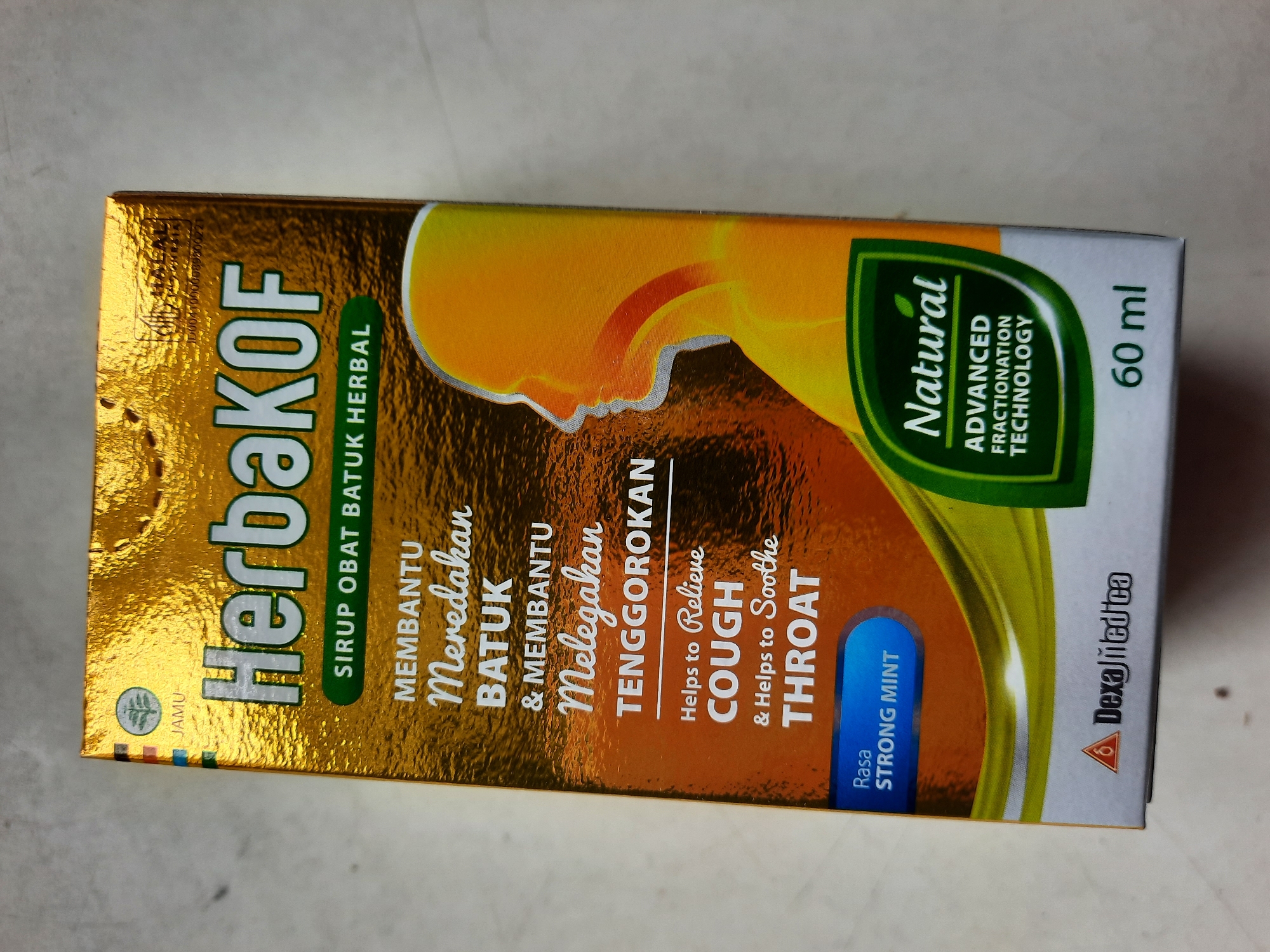 The image shows a close-up, slightly angled view of a box of herbal cough syrup. The box is predominantly gold and features text in both Indonesian and English, indicating it is for cough and throat relief. The Indonesian text reads "HerbaKOF SIRUP OBAT BATUK HERBAL MEMBANTU Meredakan BATUK & MEMBANTU Melegakan TENGGOROKAN," and the English translation reads "Helps to Relieve COUGH & Helps to Soothe THROAT." A stylized graphic of a human head and throat is present on the right side of the box, highlighting the area of relief. A green leaf-shaped emblem on the box states "Natural ADVANCED FRACTIONATION TECHNOLOGY." There is also text indicating "Rasa STRONG MINT" and a volume of "60 ml." The Dexa-Medica logo is visible in the bottom left corner. The box is resting on a light-colored, textured surface. The background is out of focus, suggesting the image was taken indoors, likely in a retail or home environment. There is no indication of Lhokseumawe, Indonesia within the visible elements of the image.