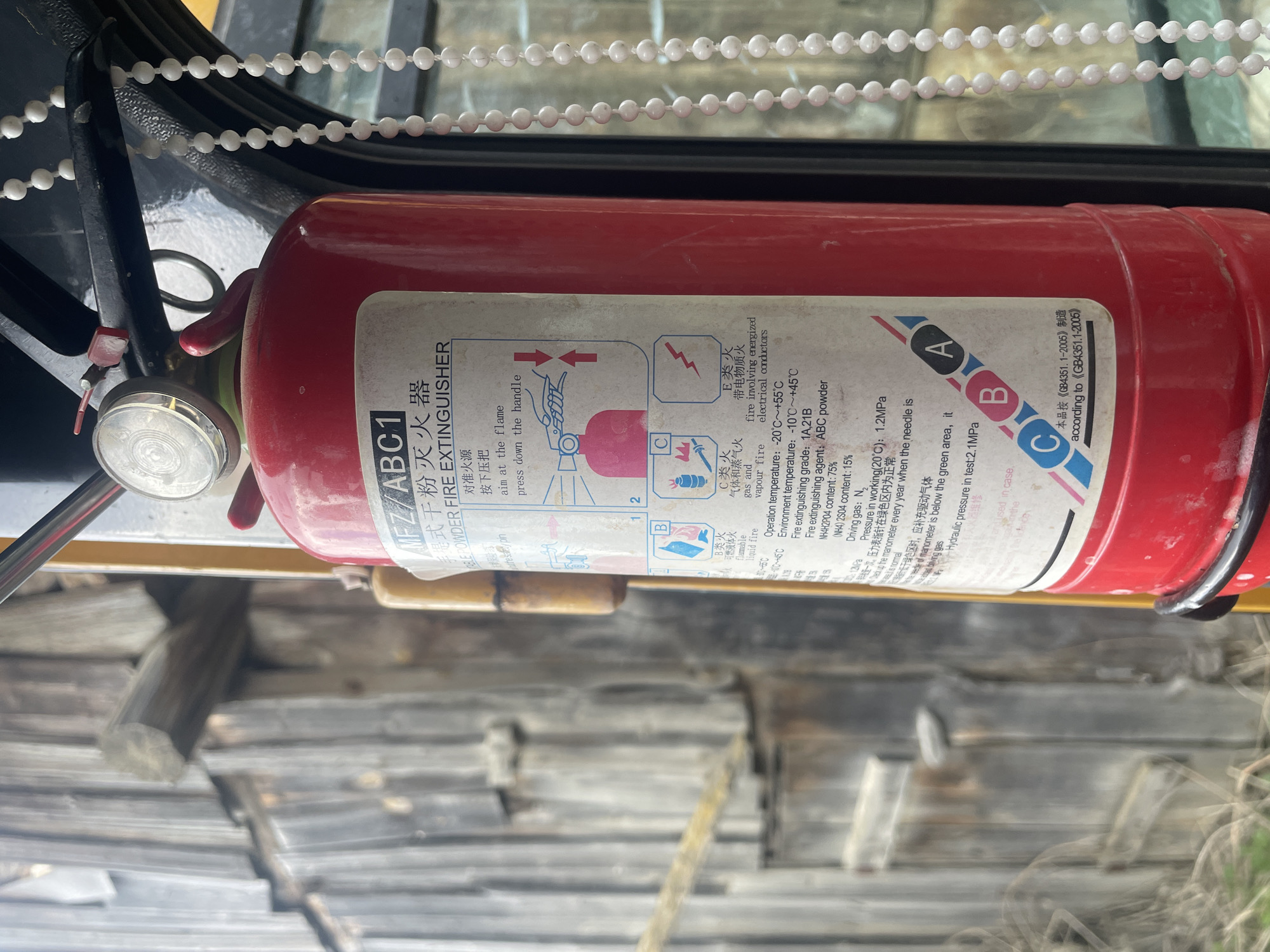 The image depicts a red MFZ/ABC1 dry powder fire extinguisher. The label on the extinguisher includes instructions for use (aim at the flame, press down the handle), as well as information about the types of fires it is suitable for (A, B, C, and E). Text details operation and environmental temperature ranges, fire extinguishing grade, composition of the extinguishing agent and driving gas, and working pressure. It also indicates that the product is manufactured according to GB4351.1-2005. The extinguisher is positioned in front of a wooden structure. The location is not evident.