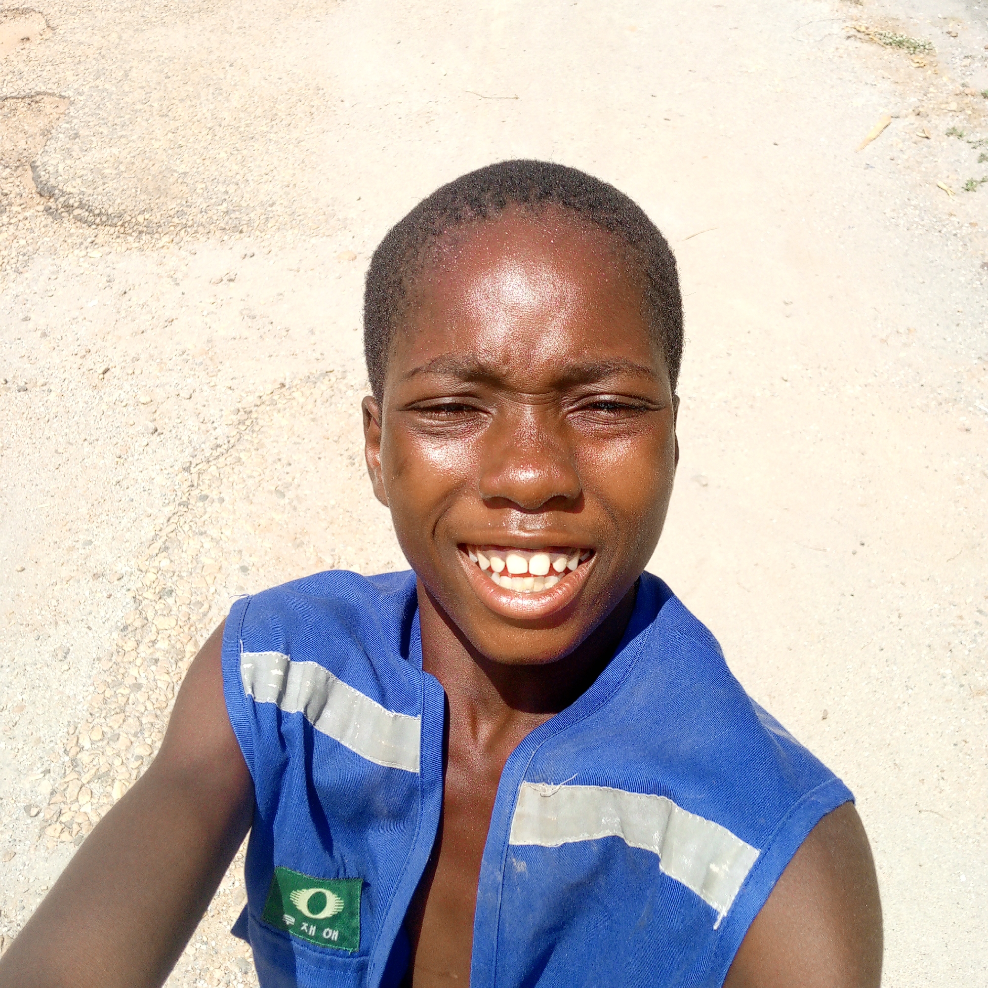 A high-angle, close-up portrait captures a young person with dark skin and short, dark hair, smiling broadly at the camera. Their eyes are crinkled from the wide smile, revealing their upper teeth. The person's forehead and cheeks appear shiny, likely from sweat or direct sunlight, reflecting the bright light.
They are wearing a blue sleeveless garment, possibly a vest or work uniform, which features two prominent horizontal reflective grey stripes across the chest area. On the lower left side of the garment, there is a green patch. This patch displays a white circular logo that resembles an eye or the letter "O" with two curved lines. Below the logo, Korean characters are visible, which appear to spell "재 애".
The background consists of light-colored, unpaved ground, characterized by small stones, dirt, and gravel, indicating an outdoor setting. The strong, direct lighting suggests it is daytime, possibly midday, with clear skies. The subject's bright smile conveys an emotion of happiness or cheerfulness.