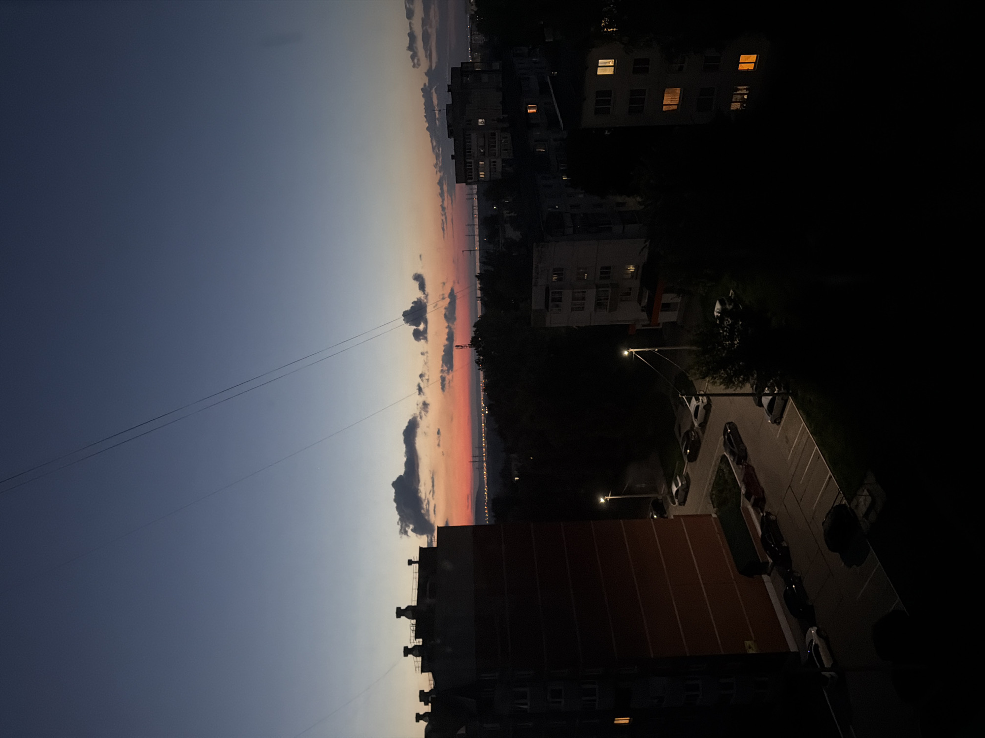 The image captures a twilight view of an urban landscape, likely in Chaikovsky, Russia. The sky transitions from a deep blue at the top to shades of orange and pink near the horizon, indicating the end of the day. A few clouds are scattered across the sky. In the foreground, on the left, is a tall, multi-story building with red brick detailing. Several cars are parked in a lot in front of this building. To the right, other buildings of varying heights and architectural styles are visible, some with lit windows. Trees and foliage are interspersed between the buildings, casting dark shadows. Power lines crisscross the sky. The overall scene depicts a residential area at dusk, with the artificial lights of buildings beginning to illuminate the darkening surroundings. There are no people visible in the image.