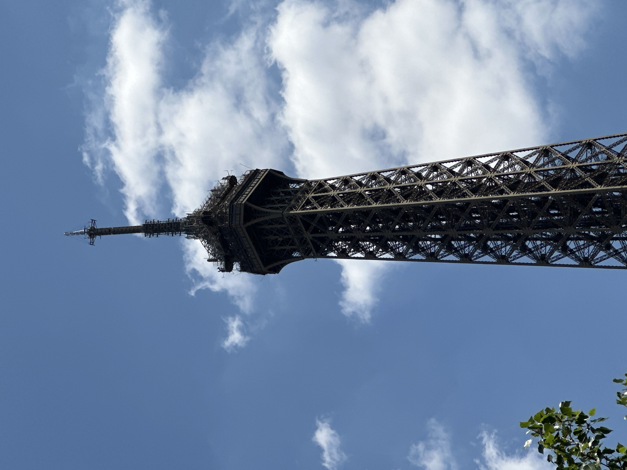 The image shows a low-angle, close-up view of the upper section of the Eiffel Tower against a bright blue sky with scattered white clouds. The intricate lattice structure of the tower is visible, along with the platforms and antennas at its summit. Some scaffolding or maintenance equipment is present near the top. In the lower left corner, a few green leaves from a tree are visible, providing a contrast to the metallic structure and the sky. The scene appears to be set in Paris, France, given the iconic nature of the Eiffel Tower. There are no people clearly visible in the frame. The lighting suggests it is daytime, likely sunny.