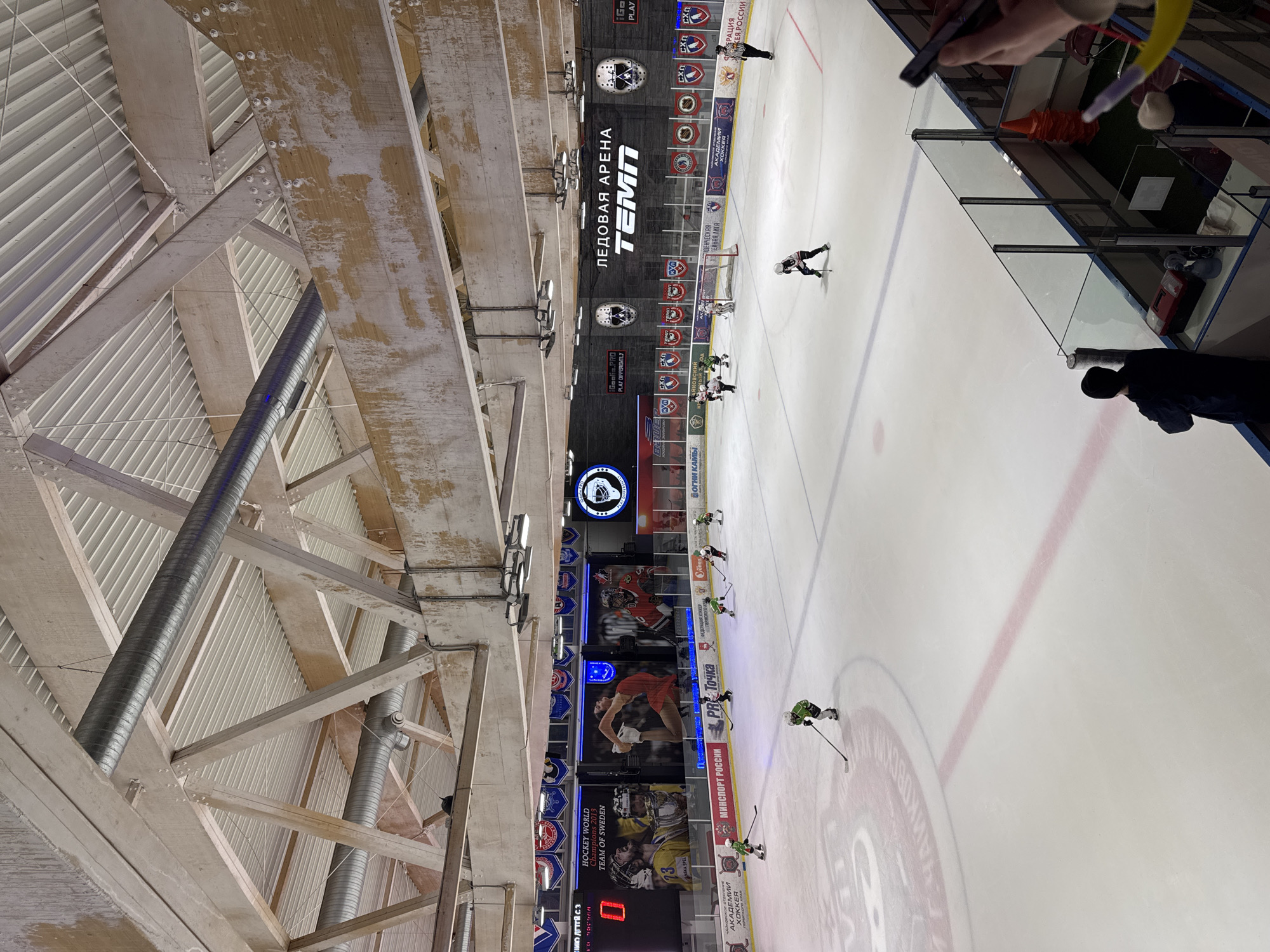The image captures an indoor hockey arena in Chaikovsky, Russia, likely during a game. Players in green and white uniforms are visible on the ice, alongside an official in a black and white striped uniform. The boards surrounding the rink are adorned with logos and advertisements, including the text "ЛЕДОВАЯ АРЕНА TEMA" which translates to "Ice Arena TEMA". Above the ice, a large video screen displays various graphics and advertisements. The arena's ceiling features exposed wooden beams and ductwork. Spectators are standing behind the rink boards near the side of the image.