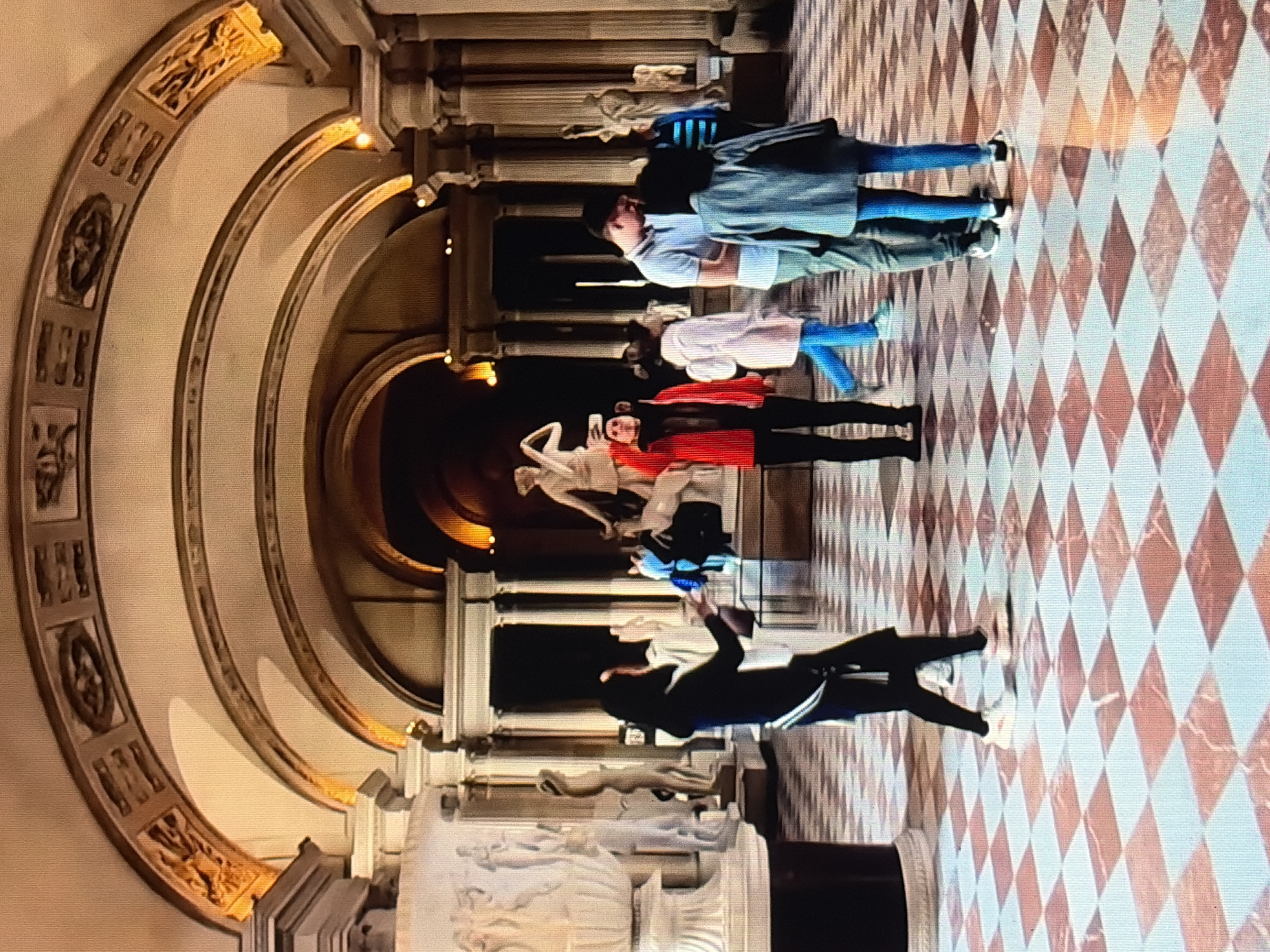 The image depicts an interior scene of a museum or art gallery, with several people viewing classical sculptures and architectural features. The environment is characterized by an arched ceiling with ornate decorations, marble columns, and a patterned floor composed of red and white diamond shapes.
In the foreground, a person wearing a black outfit with a white sash walks to the left, with their back mostly to the camera. To their right, another person in a red jacket and black pants is facing a sculpture and appears to be taking a picture or gesturing. Further into the scene, a group of people are gathered, some looking at the artwork, others conversing. One person in a light blue shirt and green pants stands with their head bowed, looking at someone in a grey jacket. Another person in a white coat walks away from the camera. In the background, more statues and architectural elements are visible, illuminated by soft lighting. The overall atmosphere suggests a quiet and contemplative experience of art appreciation.