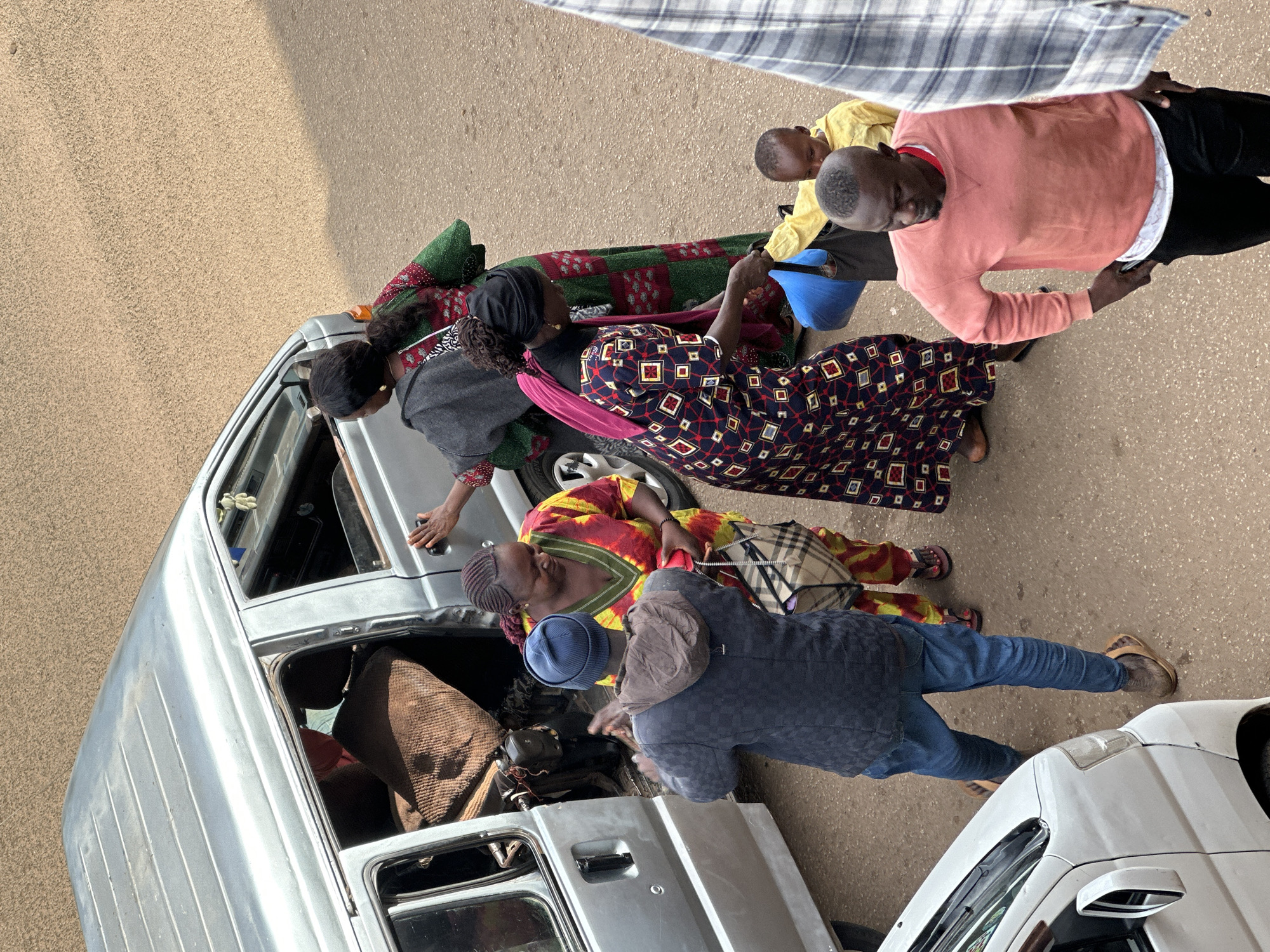 In the city of Bukuru, Nigeria, a group of individuals are gathered around an open-doored, silver minivan. A woman in a red and yellow patterned dress stands near the van, holding a checkered bag. She appears to be interacting with a man wearing a dark blue checkered jacket and a blue beanie. The man is facing away from the camera, with his hands clasped in front of him.
To the right of the minivan, a woman in a dark headscarf and a long, patterned dress is handing a blue bag to a young child wearing a yellow shirt. Another woman in a red and green patterned dress stands behind them, with her arm extended towards the minivan door. In the foreground, a man in a salmon-colored sweatshirt and black pants stands with his hands on his hips, looking downwards. The ground is a rough, sandy surface, characteristic of a Nigerian street. A portion of a white vehicle is visible on the left edge of the image, and a section of a person wearing a plaid shirt is on the right.