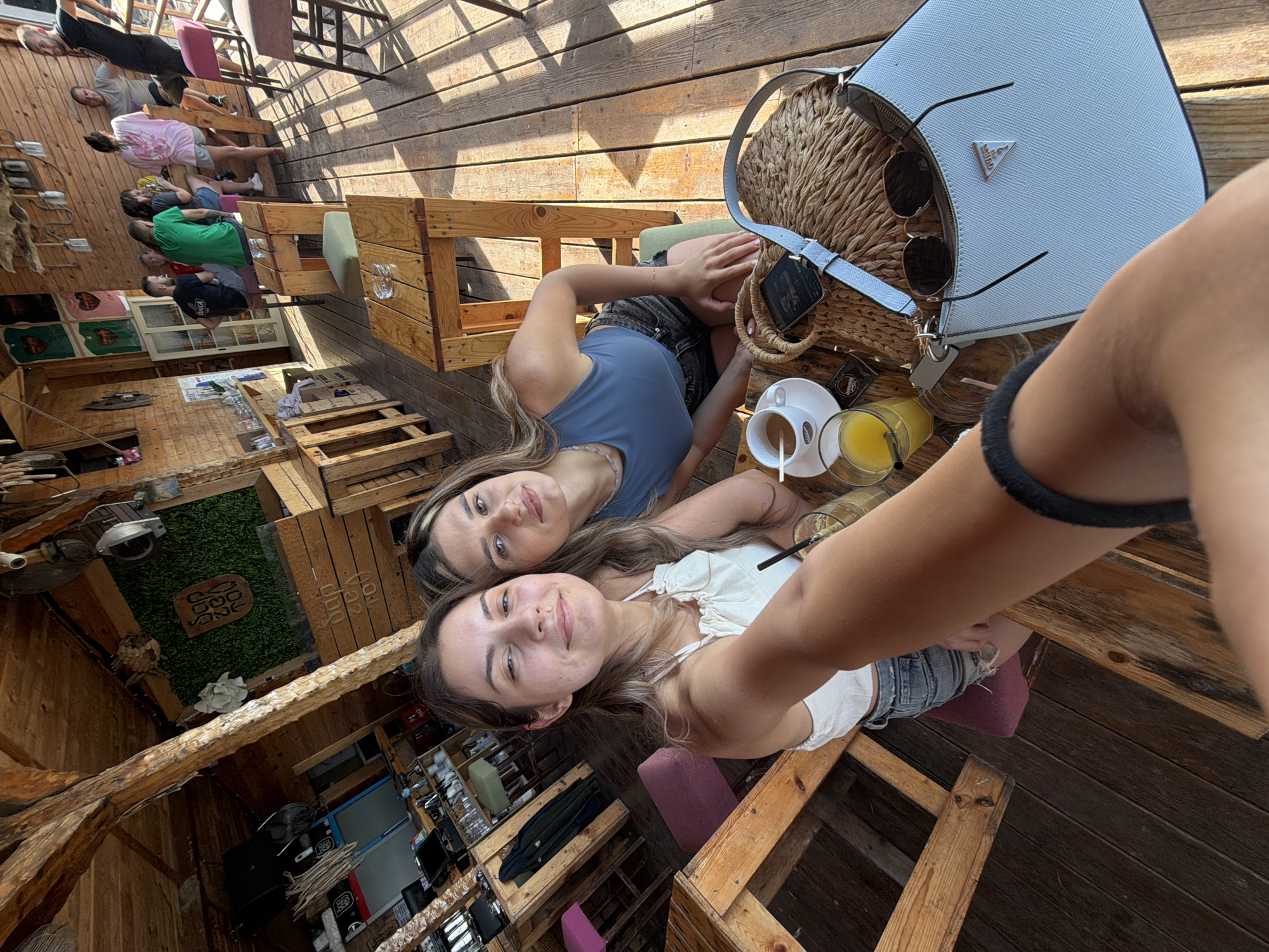 The image depicts a group of people enjoying themselves at an open-air bar or cafe in Lozenets, Bulgaria. The setting appears rustic, with wooden structures and furniture, characteristic of a beachside establishment. In the foreground, two young women are taking a selfie, with one woman in a white top and the other in a blue top. They are seated at a wooden table where drinks and a straw bag are visible. Behind them, other patrons are seated at the bar and tables, engaged in conversation or enjoying their refreshments. The overall atmosphere suggests a relaxed and social gathering.