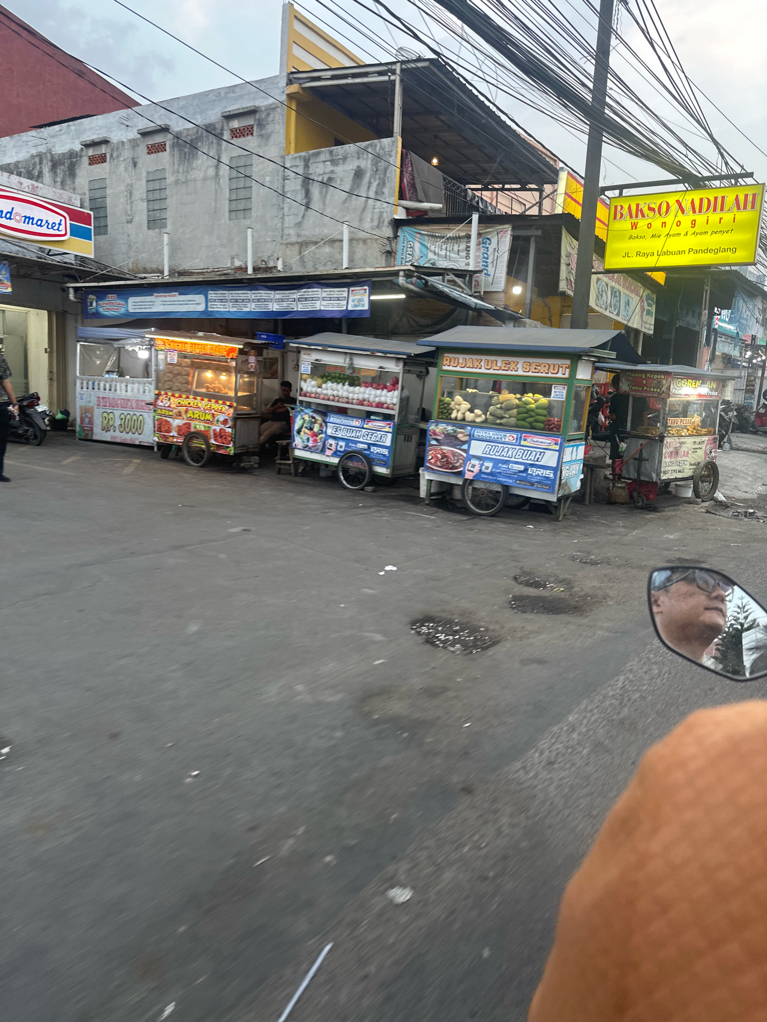The image shows a street scene in Saruni, Indonesia, with several food carts lined up along the road. The carts sell various items, including fried chicken, fruit salad, and soup. There are also several buildings visible in the background, including a yellow and gray building with a sign that reads "BAKSO NADILAH". The sky is overcast, and the road is wet, suggesting it may have recently rained. A person's hand and shoulder are visible in the foreground, indicating the photo was taken from the perspective of someone riding a motorcycle.