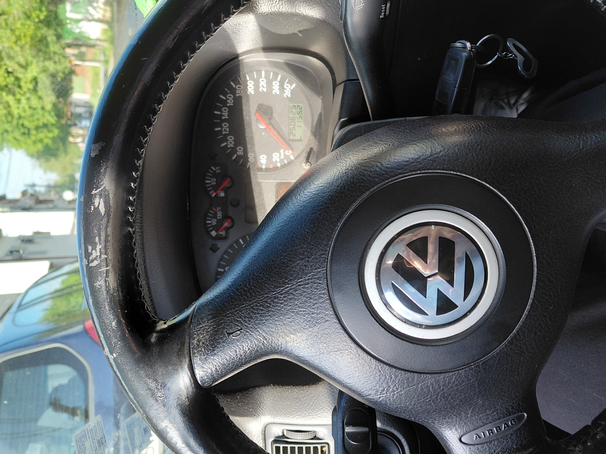 The image depicts the interior of a Volkswagen car, specifically focusing on the steering wheel and dashboard. The steering wheel, adorned with the VW logo, shows signs of wear, with peeling material along its rim. The dashboard reveals a speedometer indicating approximately 70 km/h and an odometer reading of 250625 km. Keys are inserted in the ignition. The reflection in the windshield reveals a glimpse of the outside, showing trees and a blue car. The location in Harmanli, Bulgaria, is not determinable from the image.