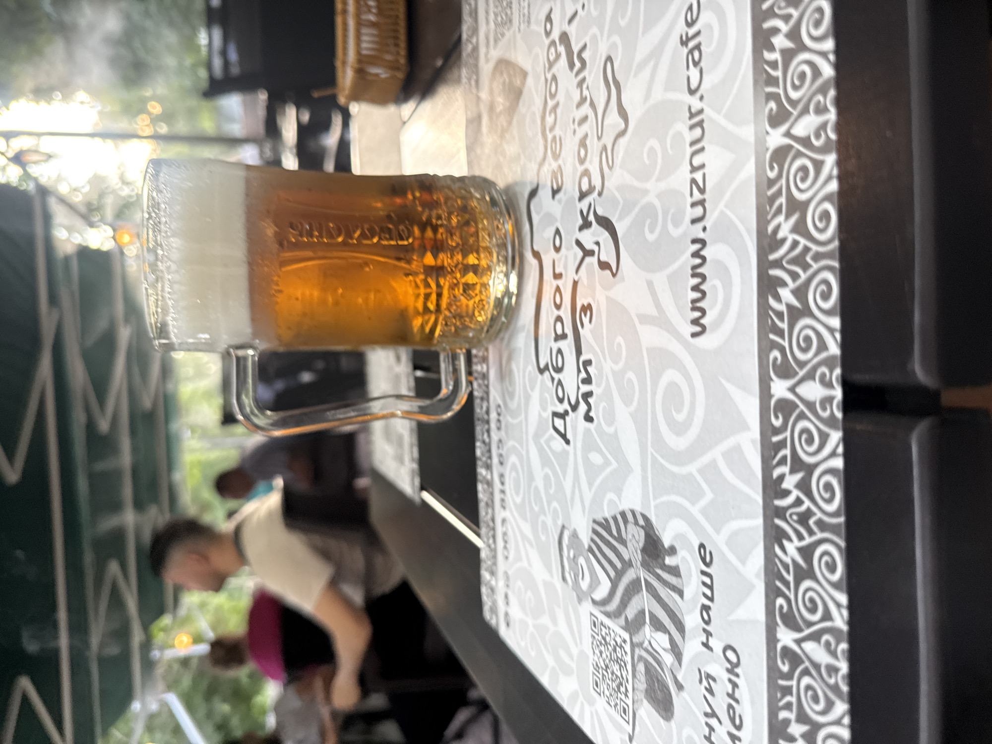 A man sits at an outdoor cafe table with a full glass of beer. The table is covered with a menu featuring a QR code and a graphic of a smiling man in traditional clothing. The menu also displays the website "www.uznur.cafe" and text in Ukrainian. In the background, other patrons are visible, and sunlight streams through an overhead canopy. The scene appears to be in Odesa, Ukraine, given the Ukrainian text on the menu.