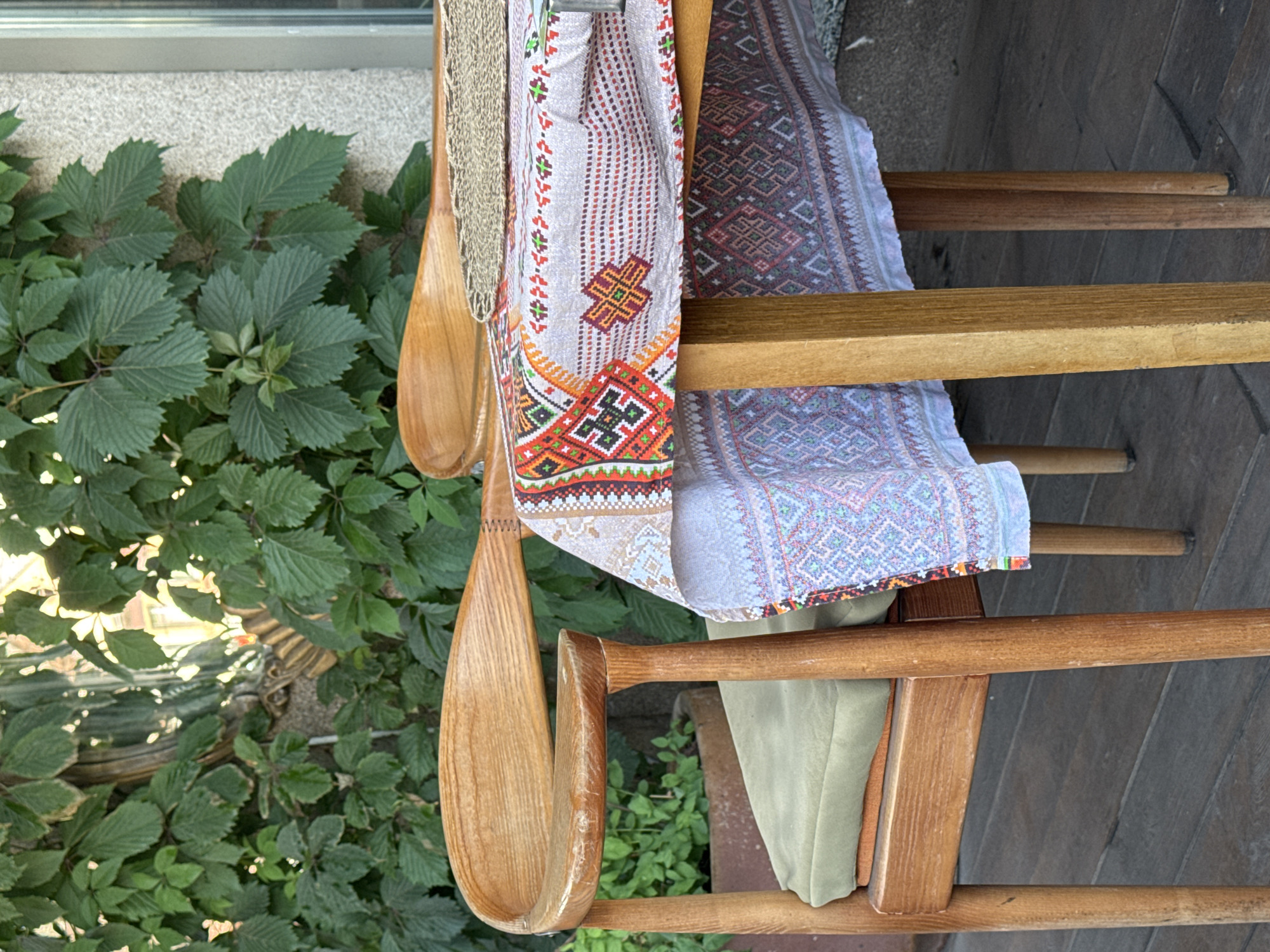 The image depicts a close-up view of a wooden chair with a green cushion, placed next to a table covered with a richly embroidered tablecloth. The tablecloth features intricate geometric patterns in red, orange, green, and blue on a light-colored fabric. A wooden serving spoon rests on the tablecloth. In the background, lush green foliage covers a wall, suggesting an outdoor or semi-outdoor setting, possibly a cafe or restaurant patio in Kryvyi Rih, Ukraine. The wooden floorboards are visible beneath the chair and table. The scene is lit by natural daylight.