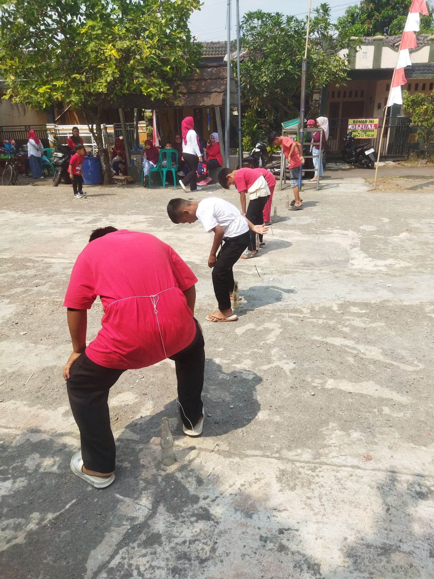 In Banjar, Indonesia, under a bright, sunny sky, a group of young people are participating in a game on a concrete courtyard. The game appears to involve knocking over glass bottles placed on the ground. In the foreground, a person wearing a bright red t-shirt and black pants is bent over, their hands resting on their knees, focused on the task. A white string is tied around their waist, connecting to something behind them. Behind them, other participants are also engaged in the game, some of them wearing white t-shirts and black pants. In the background, spectators are seated on chairs, some wearing headscarves, and a red and white Indonesian flag is visible, suggesting the event might be part of a national celebration. The environment is characterized by a paved surface, trees, and residential buildings, with a "RUMAH INI DIJUAL" (This house is for sale) sign on one of the buildings.