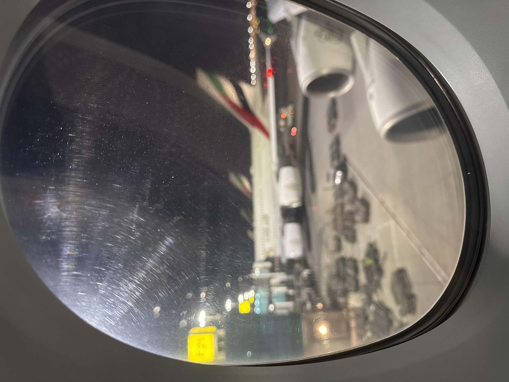The image shows a reflection in a convex mirror. The reflection depicts the interior of what appears to be a museum or exhibition space in Dubai, United Arab Emirates, based on the presence of the Emirates Airline livery on a model aircraft. There are also other models or displays within the reflection, with blurred details. The mirror's surface has visible scratches and imperfections. The background of the room is dark and it is unclear if it contains other subjects, objects, or actions.