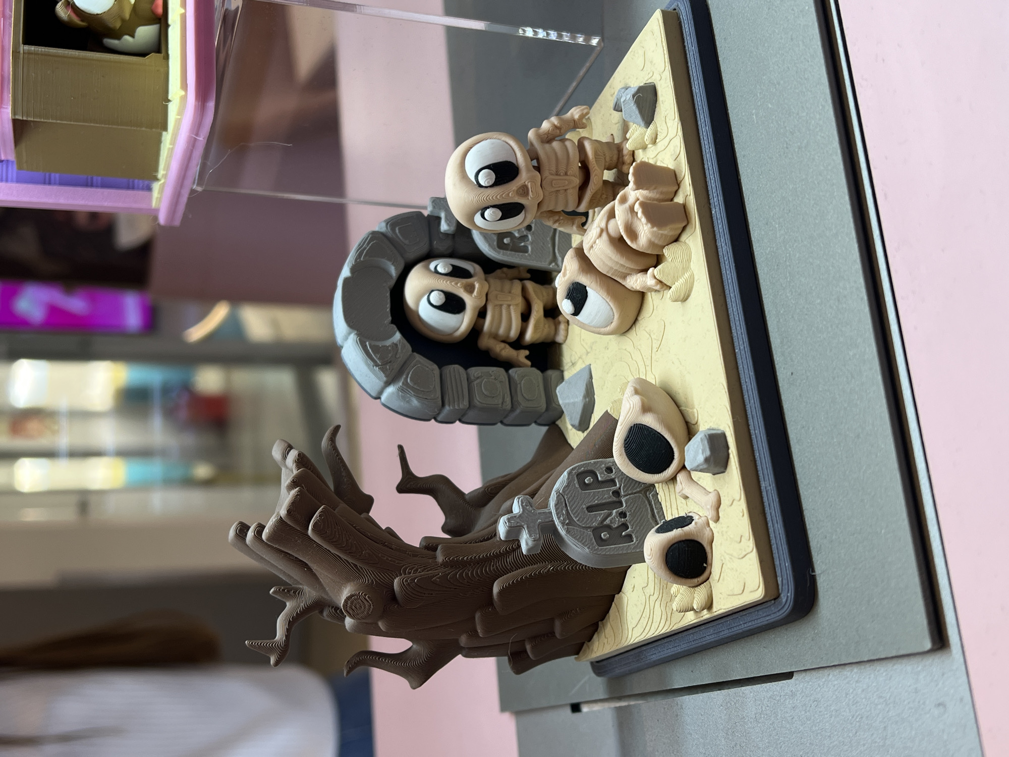 A 3D-printed diorama depicting a graveyard scene is displayed. The diorama includes two cartoonish skeletons, one emerging from a tombstone, the other lying on the beige ground. A tree-like structure with a small tombstone engraved with "R.I.P." is also visible. The entire diorama is presented within a gray frame, behind a clear display case. The background shows a blurred interior space with shelves. While the information provided does not include the diorama's exact location, the prompt provides it: Khimki, Russia.