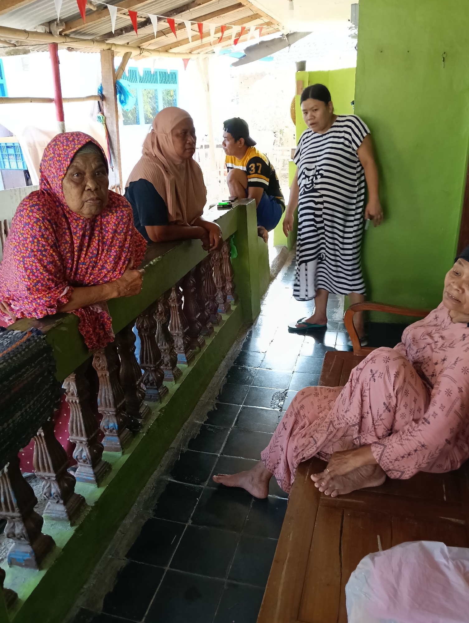 The image depicts an outdoor scene, likely in Tasikmalaya, Indonesia, where several individuals are gathered under a makeshift roof adorned with red and white pennant banners. In the foreground, an elderly woman with a patterned pink headscarf and garment is leaning on a green railing, her hands clasped. Beside her, another woman in a tan hijab and dark top also rests her hands on the railing. Further back, a young person wearing a yellow and black numbered t-shirt and a black cap is squatting. To the right, a woman in a black and white striped dress stands looking towards the viewer. In the lower right corner, another elderly woman, seated on a wooden bench and wearing a pink patterned outfit, is partially visible. The environment includes a tiled floor and a vibrant green wall.