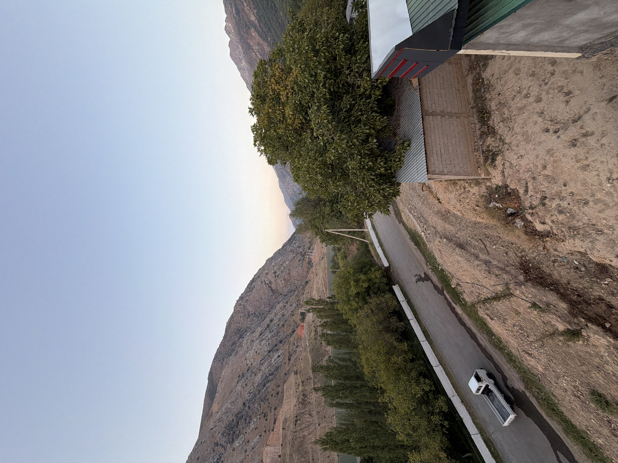 The image depicts a serene, rural landscape scene at dusk, likely in the vicinity of Nanay, Uzbekistan, as suggested by the presence of mountains and vegetation typical of the region. In the foreground, a small white pickup truck is captured in motion on a paved road. The road is flanked by a concrete barrier on one side and a steep, dusty embankment on the other, which is sparsely covered with dry grass and some scattered debris.
Further back, a dense canopy of green trees forms a verdant border along the road, suggesting a natural setting or cultivated farmland. Beyond the trees, a body of water, possibly a reservoir or river, is visible, reflecting the soft light of the evening sky. Towering mountains, characterized by their rugged, dry slopes and striated rock formations, dominate the background. The sky above is a clear, pale blue, indicative of the twilight hours.
To the right of the image, a small building with a corrugated metal roof and a brick wall is partially visible. Adjacent to it, another structure with a modern roof design featuring red accents and a green metal section is also present. The overall atmosphere is tranquil, with the fading light casting long shadows and imbuing the scene with a sense of calm. The presence of the pickup truck suggests human activity and transportation within this natural setting.