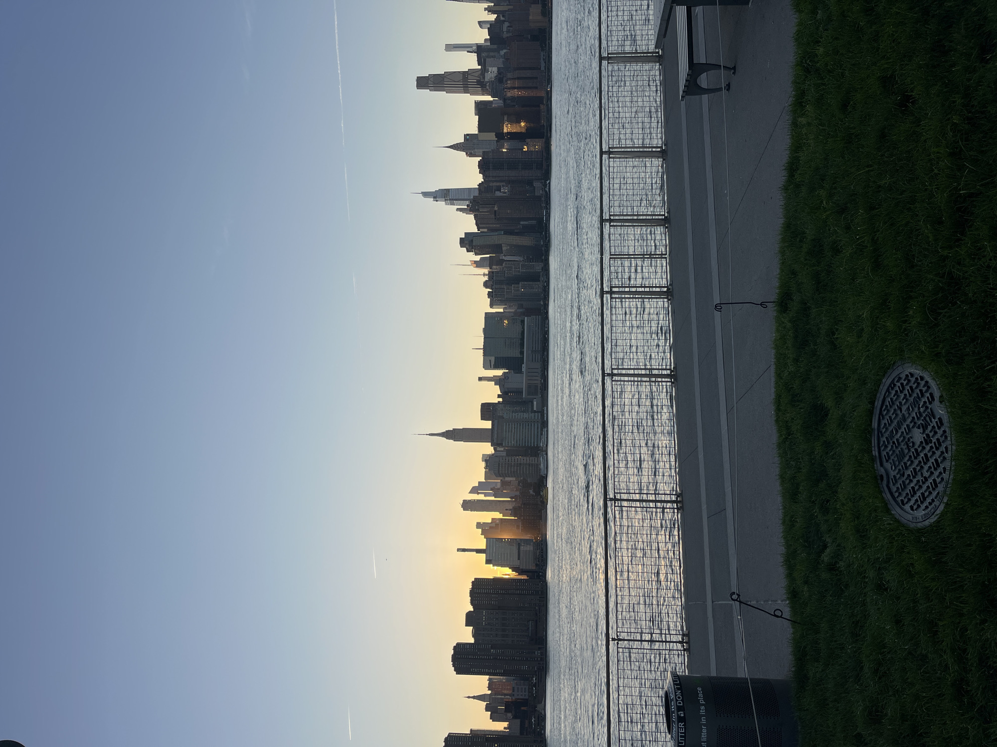 This is a wide, horizontal, eye-level photograph of the New York City skyline viewed across the East River at sunset. The sun is low on the horizon, casting a warm glow on the buildings and the water. The sky is clear and a pale blue, with a few wispy clouds. The buildings of Manhattan are silhouetted against the sky, with the Empire State Building clearly visible. The water in the foreground is choppy, with sunlight reflecting off the surface. A metal fence runs along the edge of the waterfront promenade, separating it from the grass and a manhole cover in the foreground. A dark green trash can is visible on the left side of the frame, with text on it that reads "PUT LITTER IN ITS PLACE" and "DON'T LITTER". A dark-colored bench with a metal frame is visible on the right side of the frame. The overall scene is one of urban tranquility at dusk.