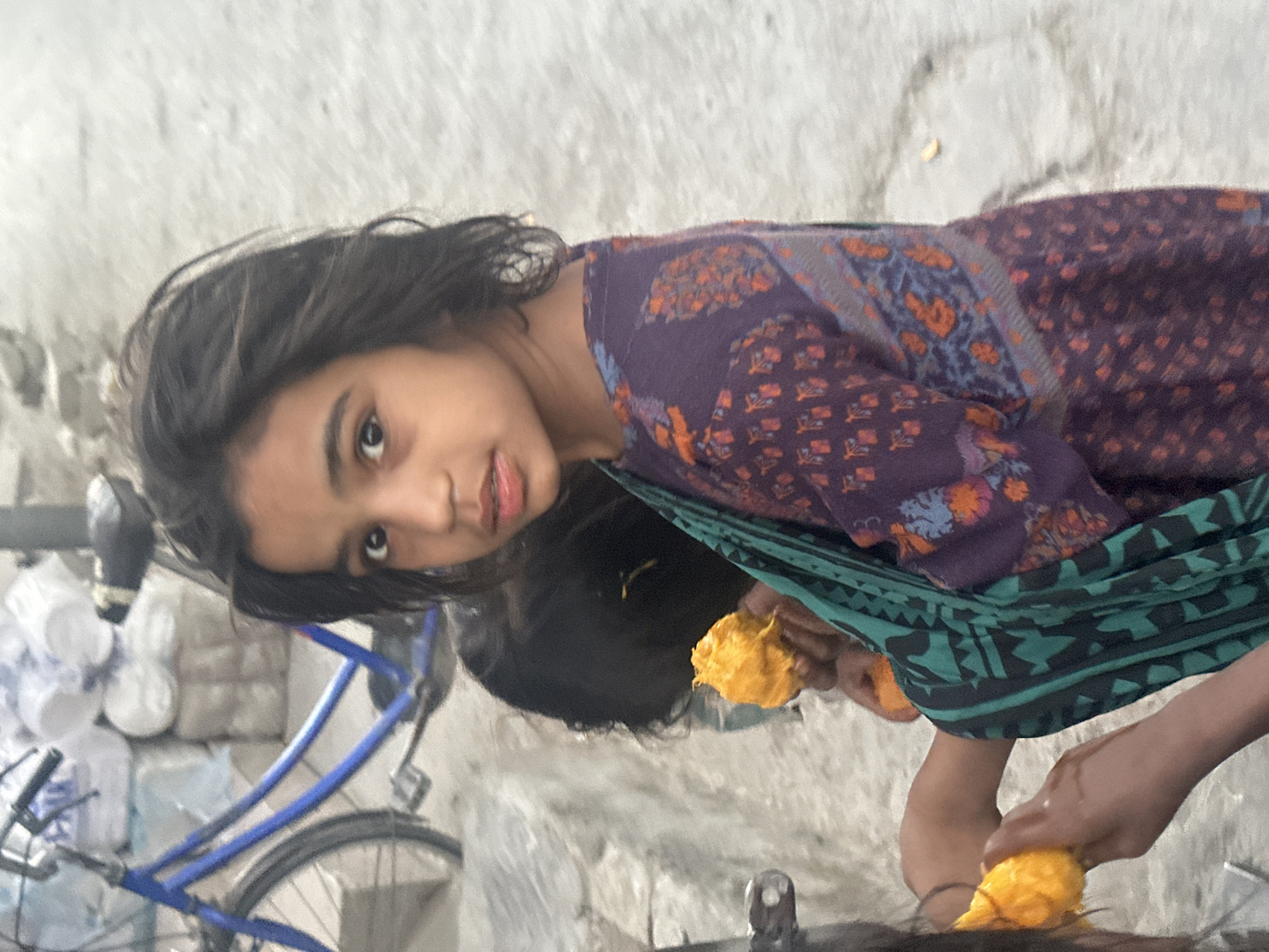The image, captured in an outdoor urban setting, shows two young girls and a bicycle. The girl in the foreground, on the right, is facing the camera and holding a piece of fruit. She has dark hair and is wearing a purple garment with an orange and blue pattern, and a green and black patterned scarf draped over her shoulder. To her left and slightly behind her, another girl is visible from the back, also holding a piece of fruit. A blue bicycle is positioned behind the girls, with stacks of white, bagged items in the background. The ground appears to be a light-colored, rough surface.