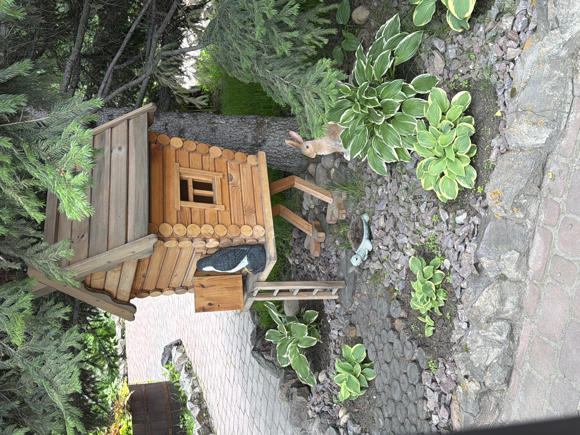 A wooden birdhouse, designed to resemble a small, rustic cabin, stands on a tree trunk in an outdoor setting. The birdhouse is constructed from round logs and features a sloped, shingled roof. A small, wooden ladder leads up to an opening on the side of the birdhouse, next to which sits a black and white statue of a bird. Below the birdhouse, a decorative garden is present. This garden includes various plants with green and white variegated leaves, several large rocks, and a pathway made of circular wooden slices. A light green, decorative turtle figurine lies on the ground near the path, and a beige, rabbit-shaped statue is positioned to the right of the birdhouse. The area is surrounded by evergreen trees, with their branches casting shadows on the scene. A paved walkway made of brick-like stones runs along the left side of the image, and a large wooden barrel is visible in the background on the left. The location is identified as Sukhaya, Russia.