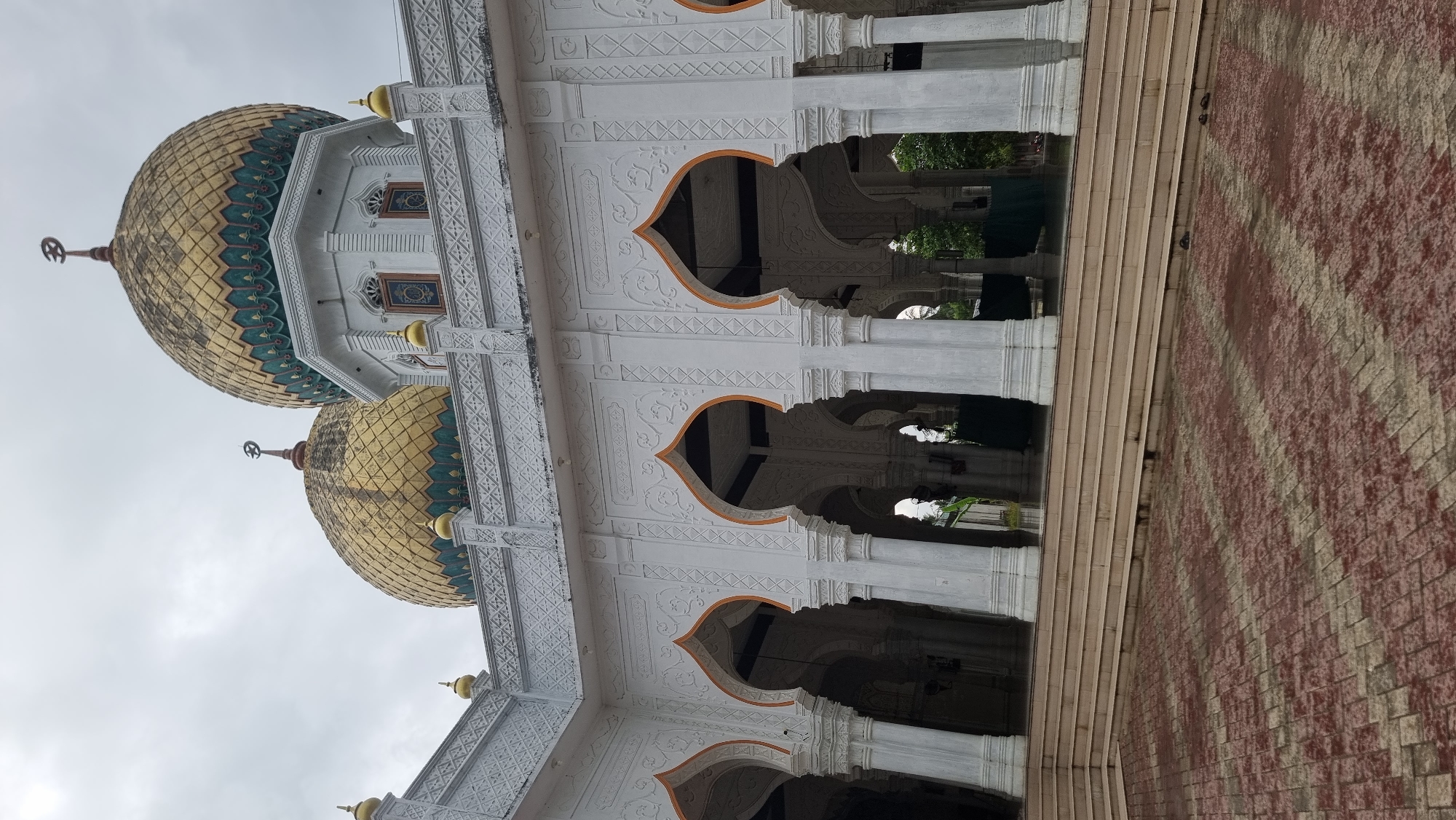 The image shows the exterior of a mosque in Meunasahlhok, Indonesia. Two prominent golden domes, intricately patterned with green and blue accents, rise against a cloudy sky. The domes are topped with crescent moon finials. The mosque features white walls with ornate relief carvings and arched colonnades. Rows of white pillars support the structure, leading into shadowed interior spaces. A set of wide, light-colored steps ascends to the colonnaded area, and beyond that, a courtyard paved with reddish-brown and beige interlocking bricks is visible. The ground appears wet, suggesting recent rain. No people are visible in the image. The overall impression is of a grand and architecturally detailed religious building.