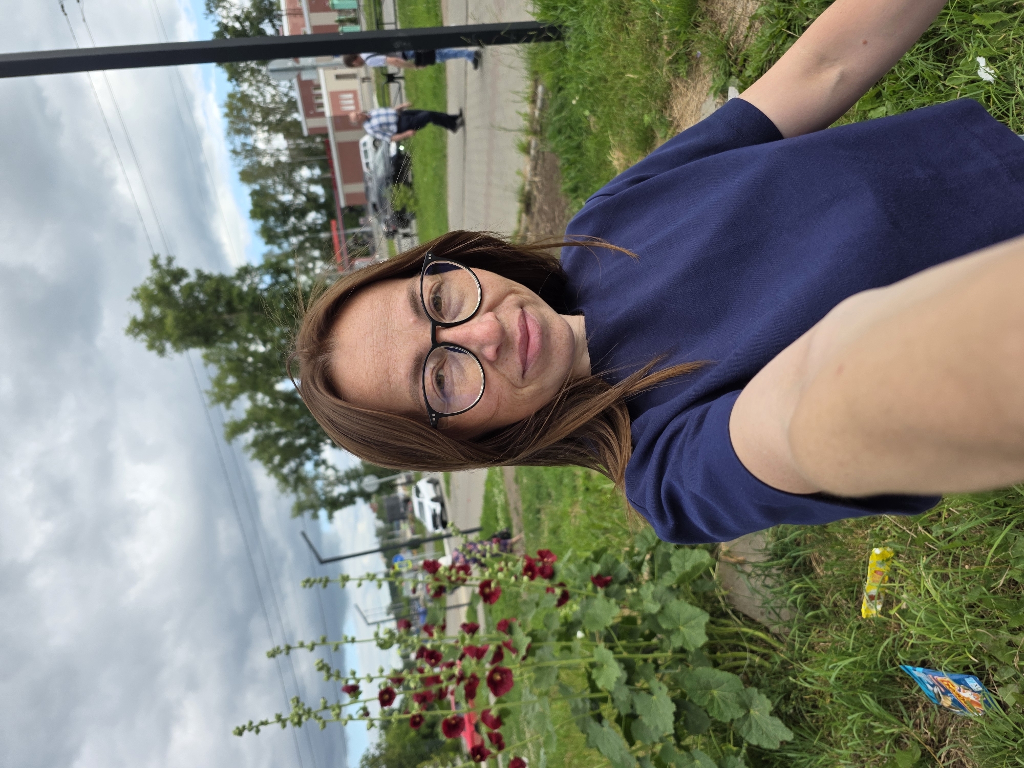 A woman with long brown hair and glasses is taking a selfie outdoors. She is wearing a navy blue t-shirt. Behind her, there are tall plants with red flowers and green leaves. In the background, there is a street with cars, buildings, and trees. The sky is cloudy. The image appears to be taken in Chaikovsky, Russia, possibly near a residential area.