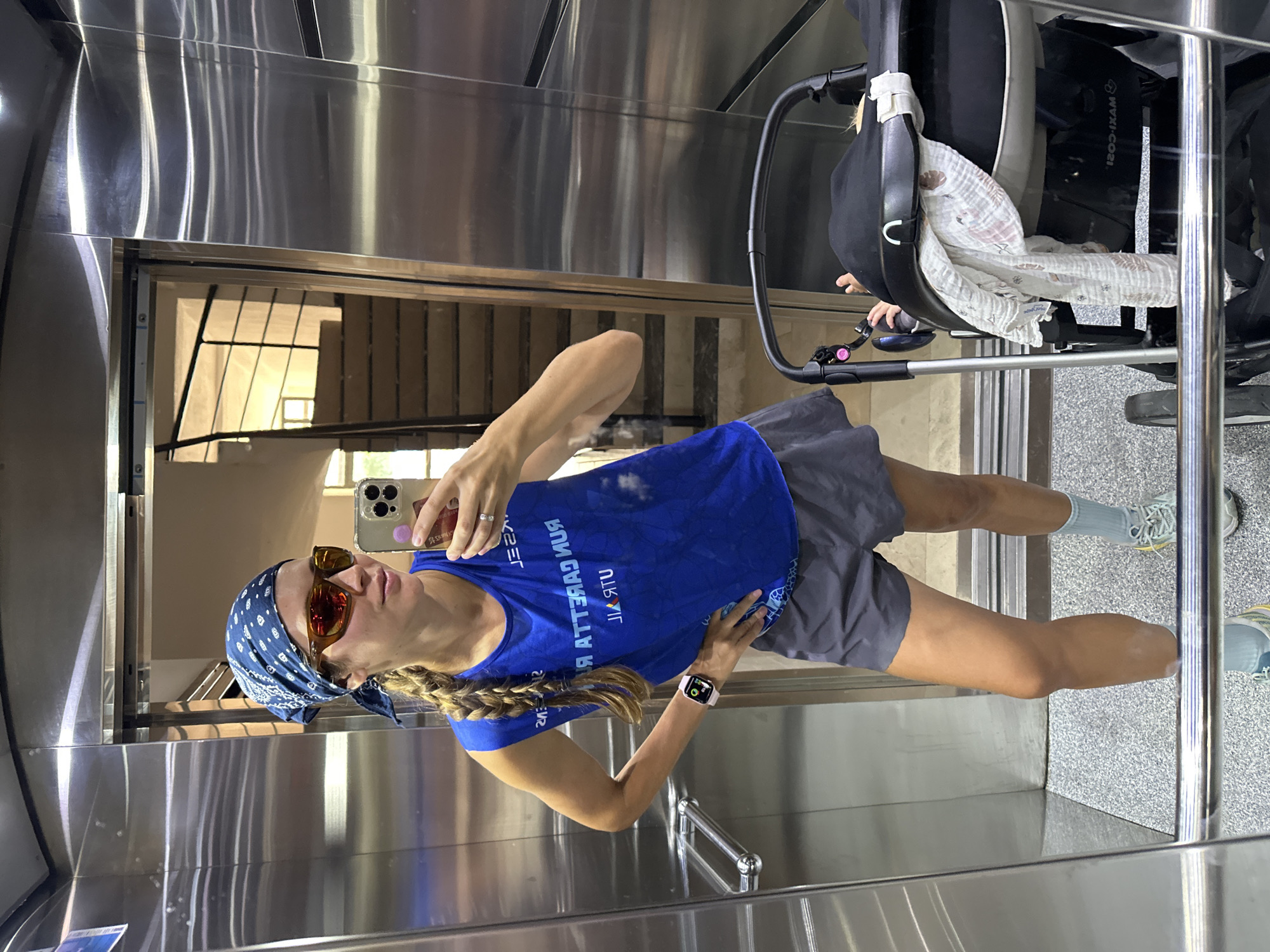 A woman is taking a mirror selfie in an elevator. She is wearing a blue tank top with white text, grey shorts, a blue bandana, sunglasses, and athletic shoes with blue and yellow accents. She has a pink watch on her left wrist. To her right, a black baby stroller with a baby covered by a blanket is visible. The elevator has mirrored walls and a speckled grey floor. In the reflection, a staircase with wooden steps and black railings is visible through an open doorway. The woman's left hand is on her hip, and she is holding her phone with her right hand to take the picture. The scene appears to be located in an apartment building or hotel, possibly in Fethiye, Turkey, given the context.